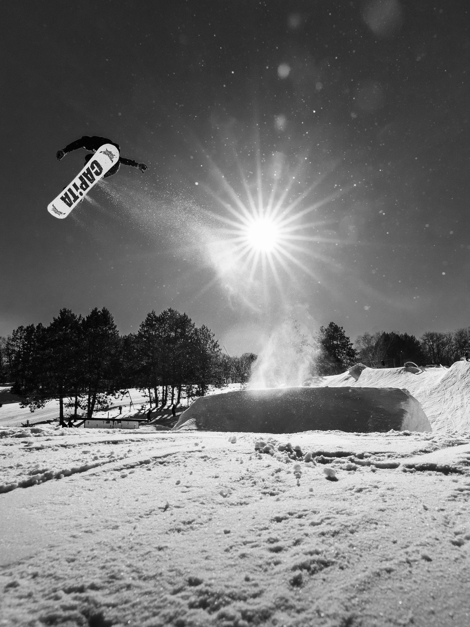 Snowboarder performing a high jump trick at a snowy terrain park on a sunny winter day, captured in black and white.