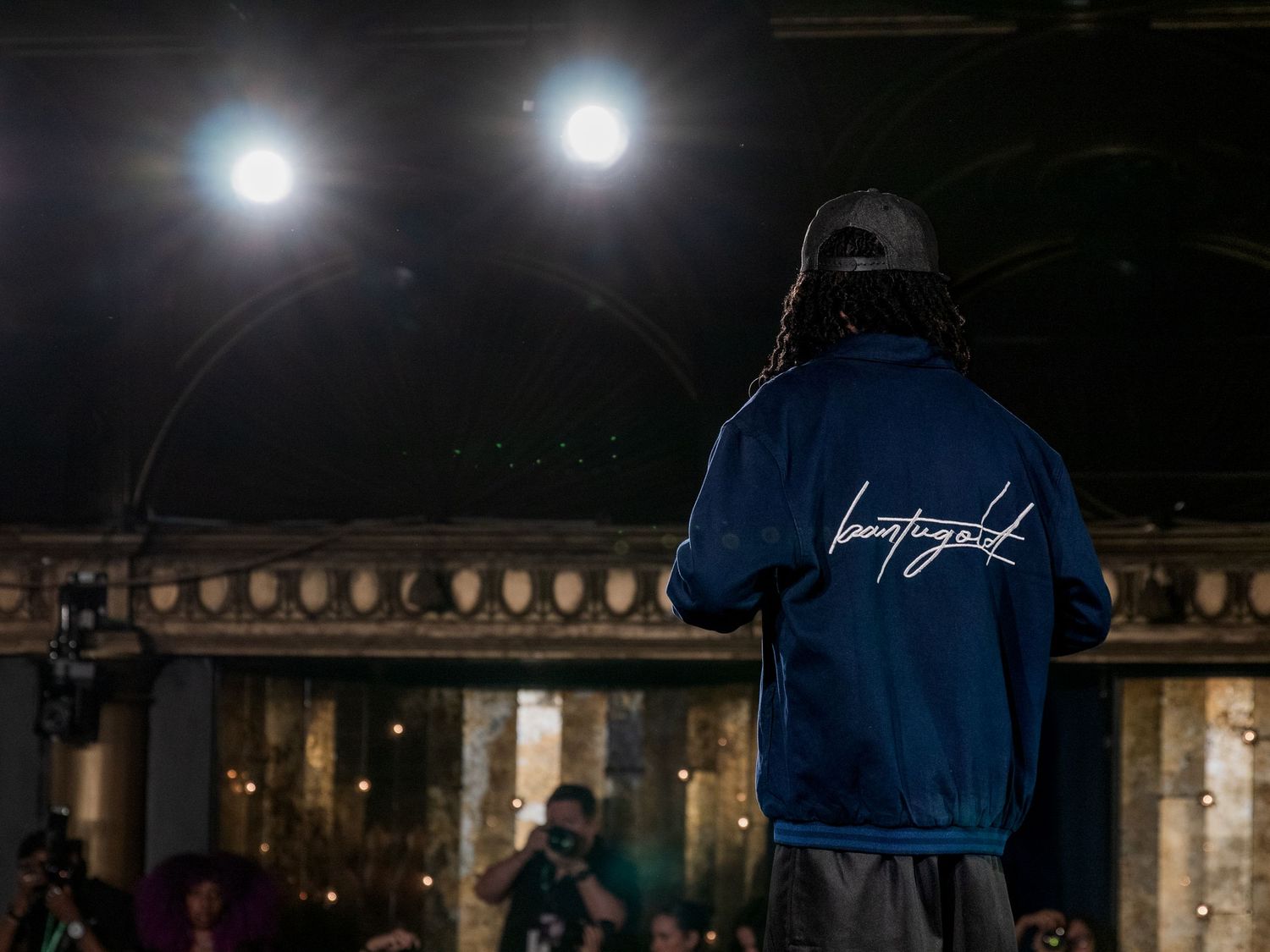 Person in blue jacket with script logo standing on dark stage under bright spotlights.