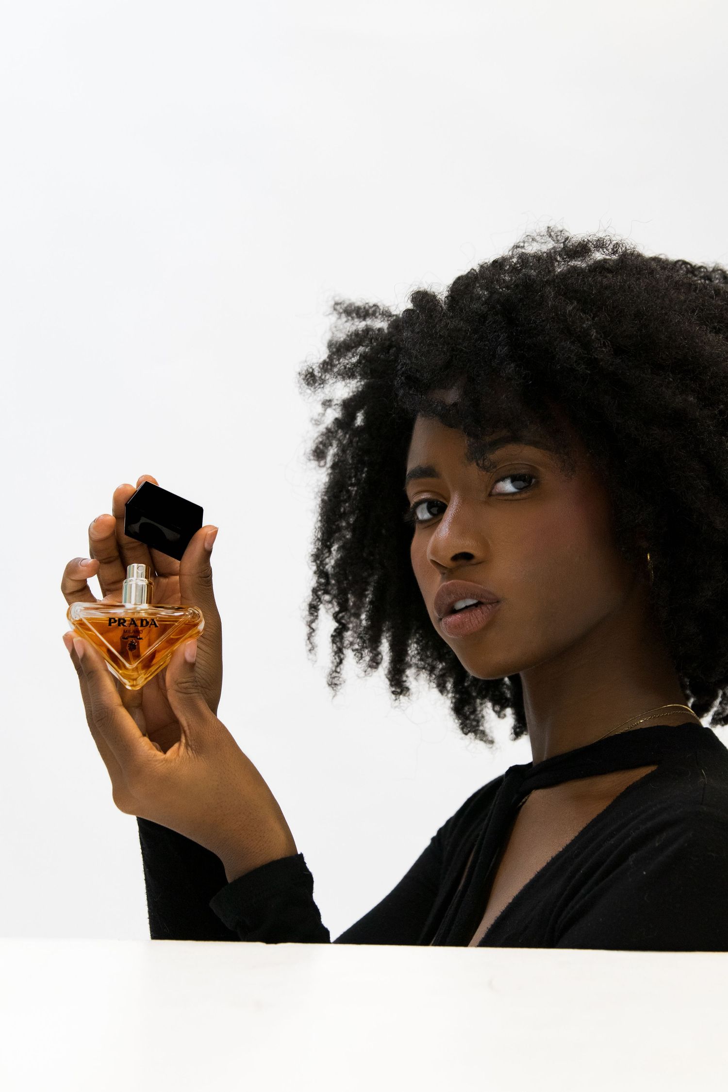 Woman with curly afro hair holding a Prada triangular perfume bottle against white background.