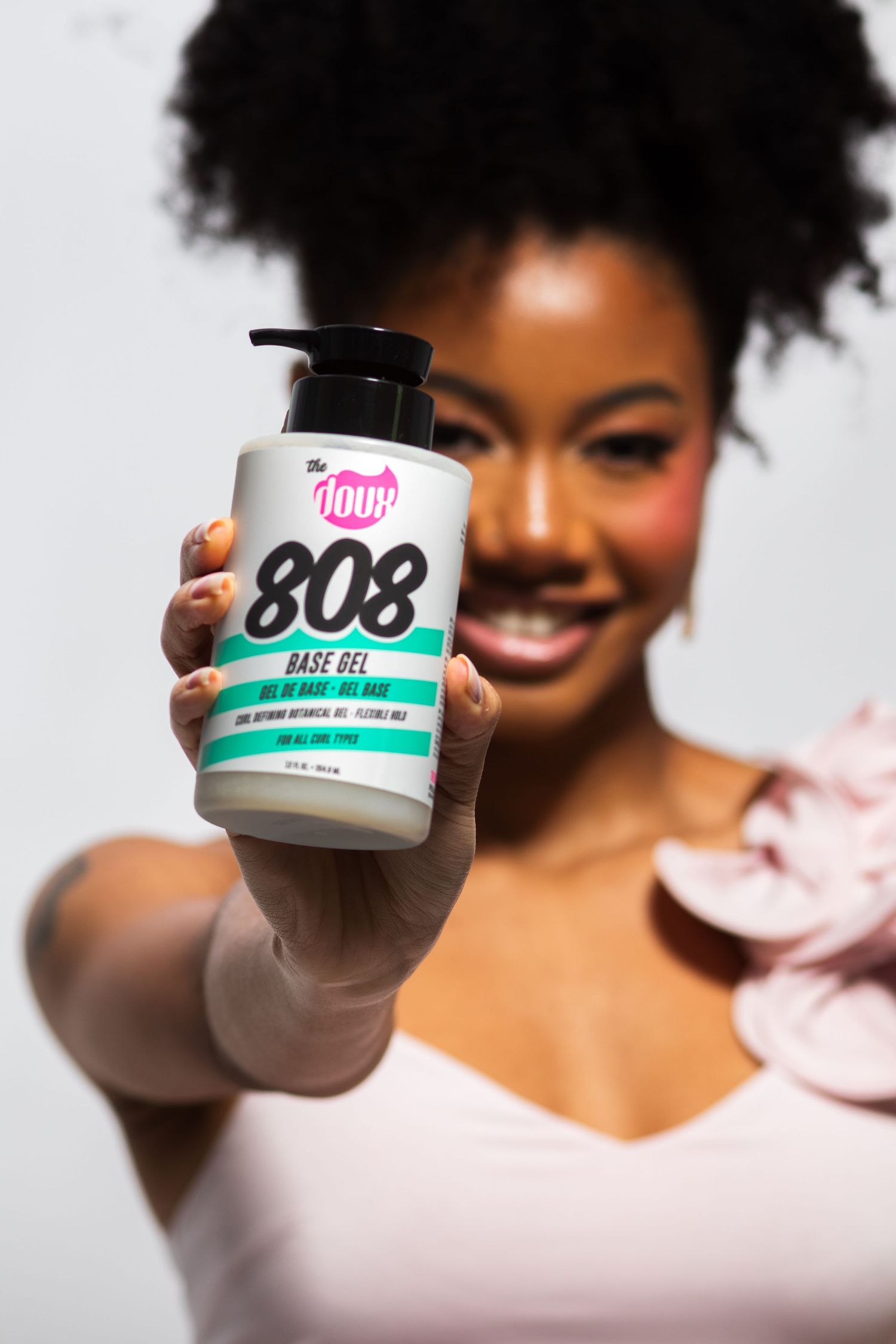 Woman with natural afro smiling while presenting The Doux 808 Base Gel pump jar forward.