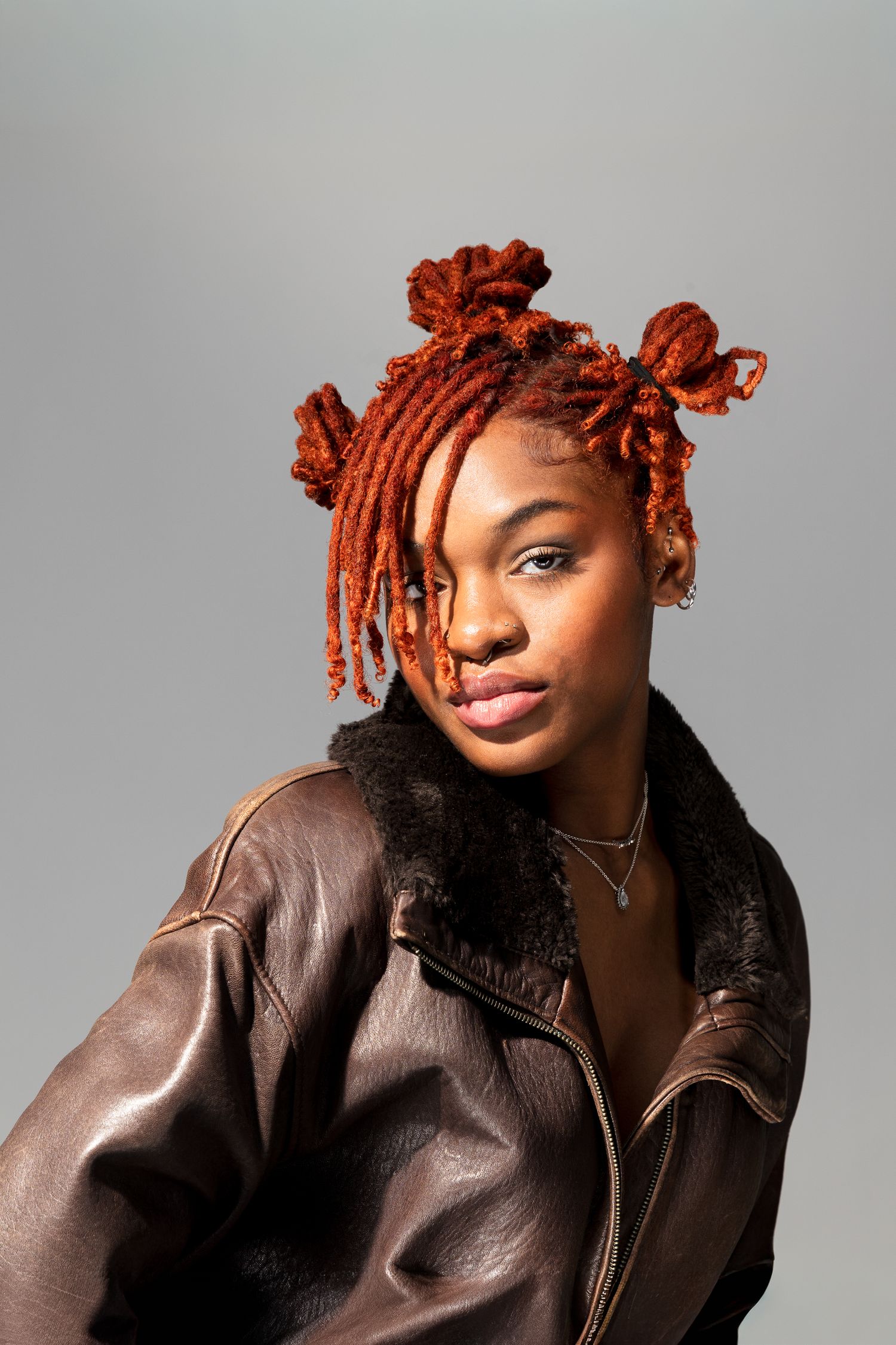 Young woman with auburn dreadlocks wearing oversized brown leather jacket poses on gray background.