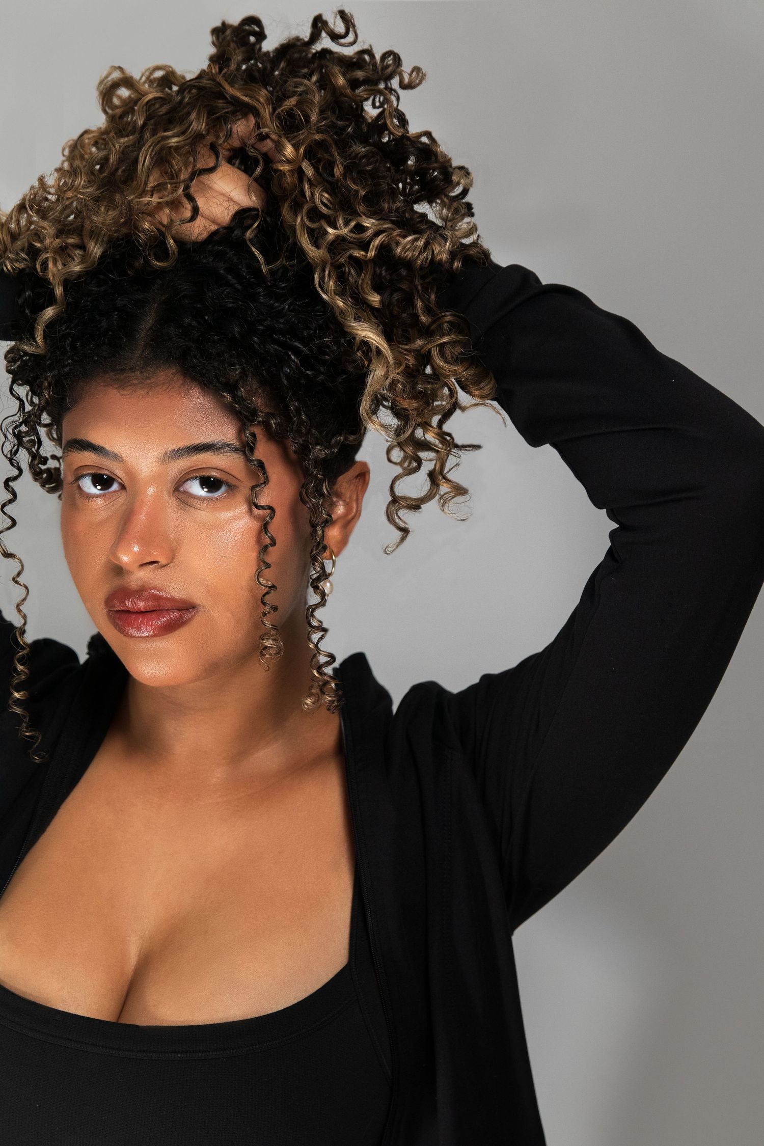 Young Black woman with highlighted curly hair posing confidently in a black outfit against a gray background.