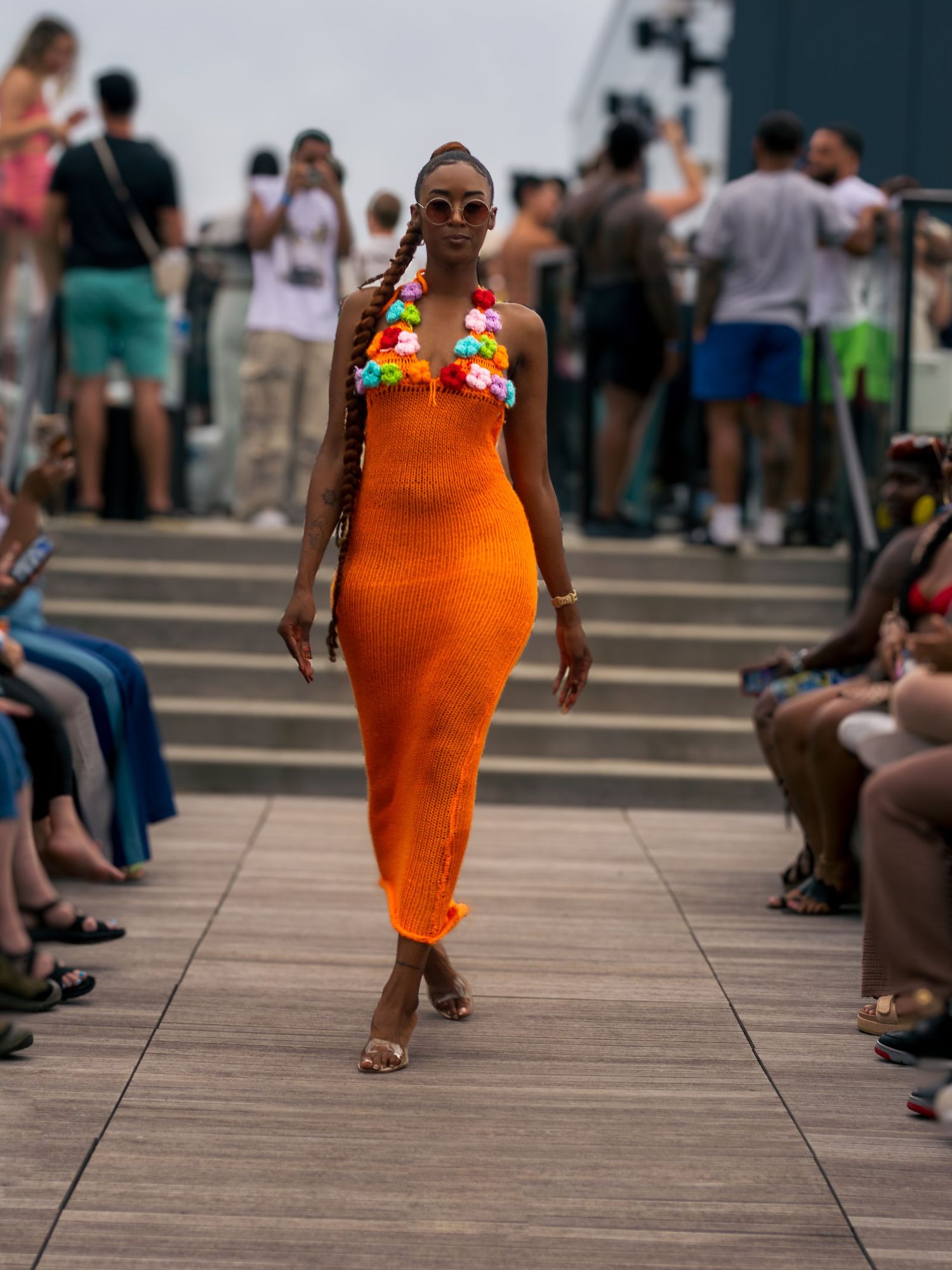 Model walks runway in orange knit midi dress with floral necklace at outdoor show.