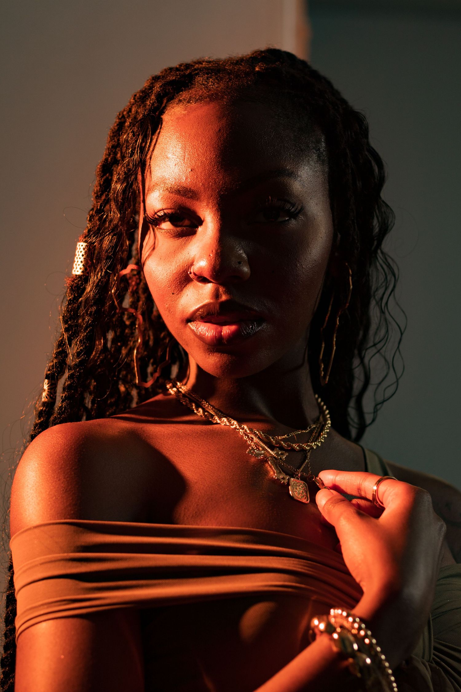 A Black woman with braids and layered gold necklaces poses in an olive off-shoulder top in warm dramatic lighting.