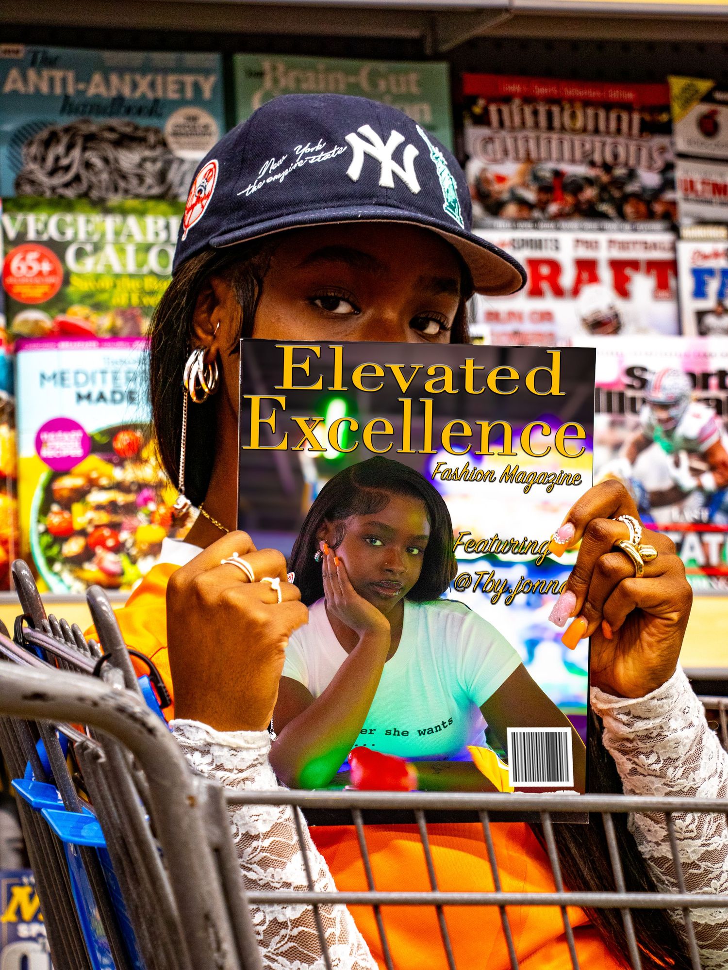 Young woman in NY Yankees cap holds Elevated Excellence Fashion Magazine in front of a newsstand display.