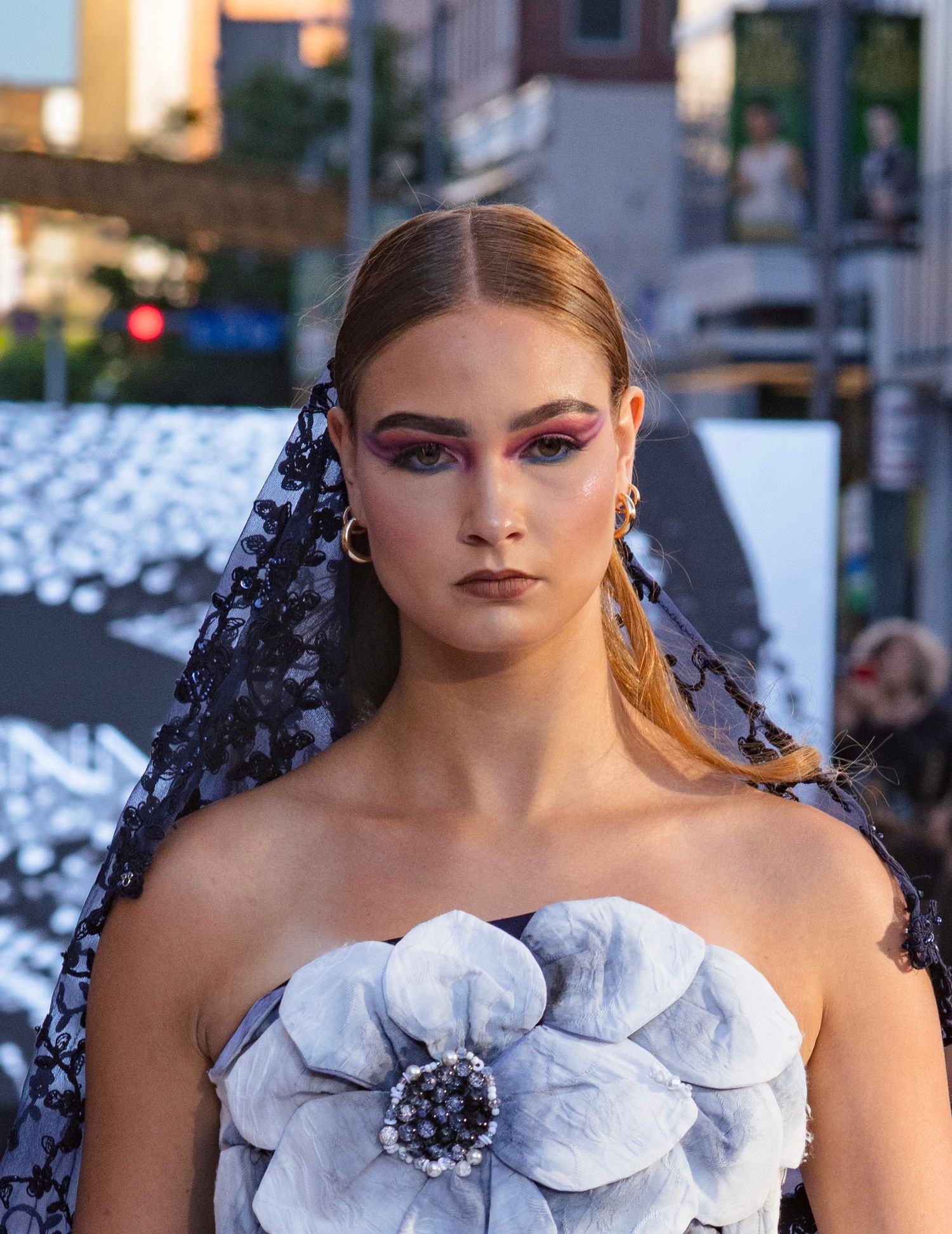 Model with bold pink eye makeup wears white floral dress and dark lace veil on runway.