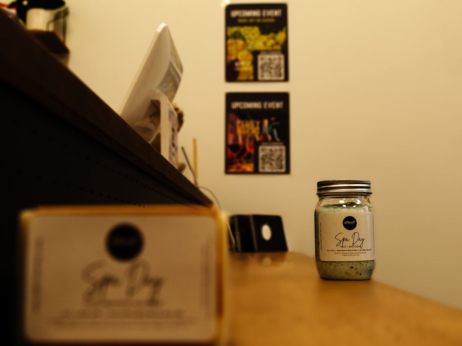 Spa Day product display on wooden counter with upcoming event flyers and QR codes on wall in background.