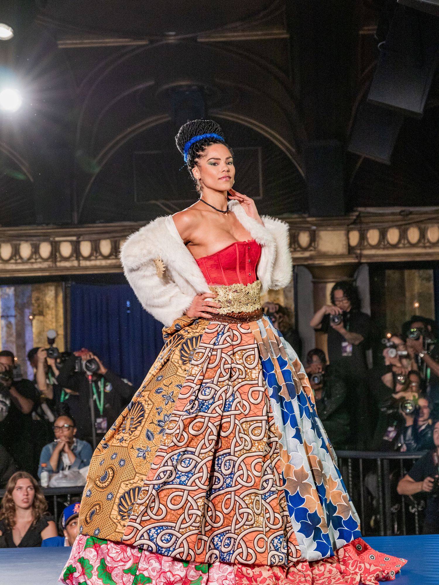 A model showcases a vibrant African print ballgown with fur wrap and blue crown headpiece on a runway.