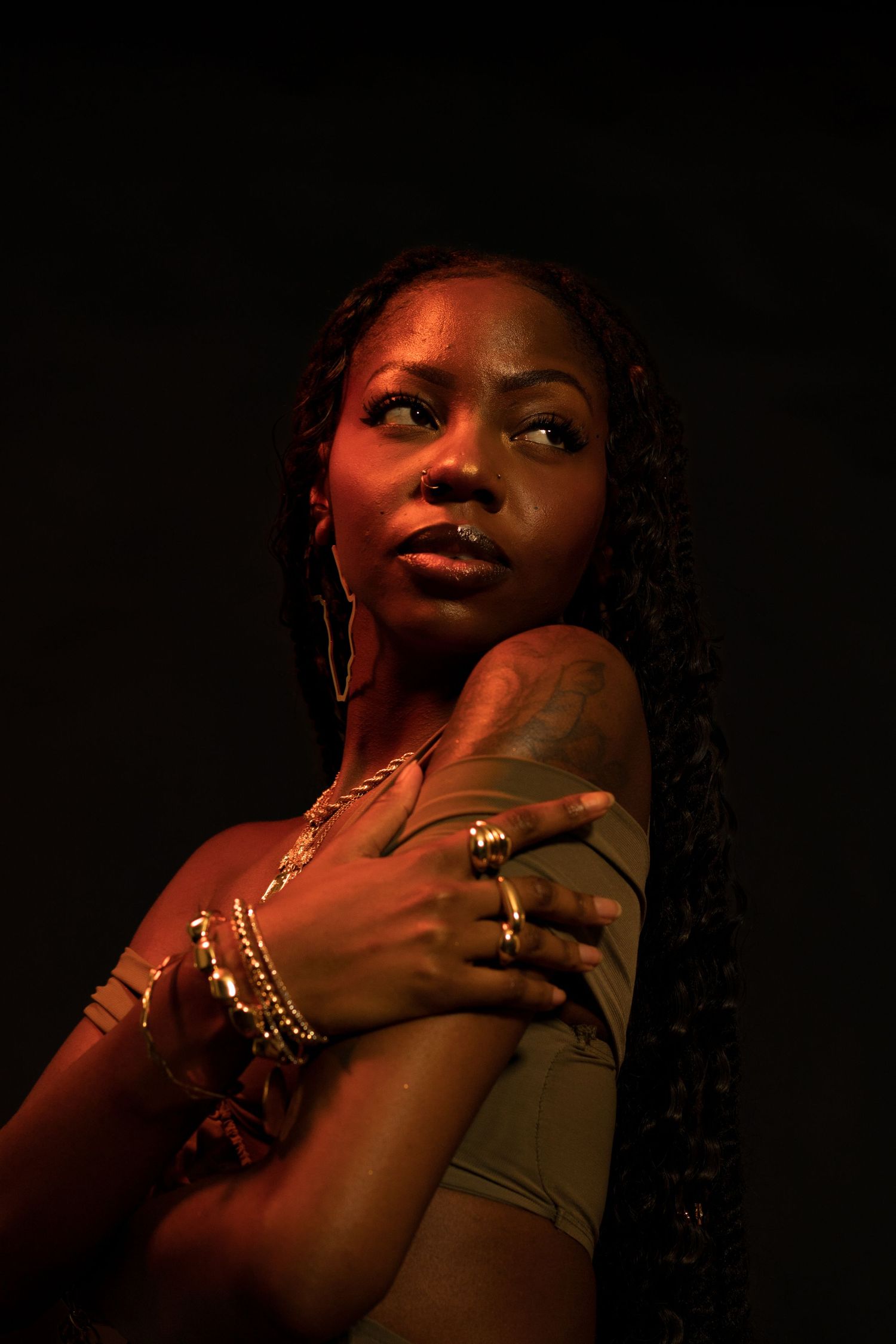 A Black woman with braids and gold jewelry crosses her arms, gazing upward in dramatic warm orange studio lighting.