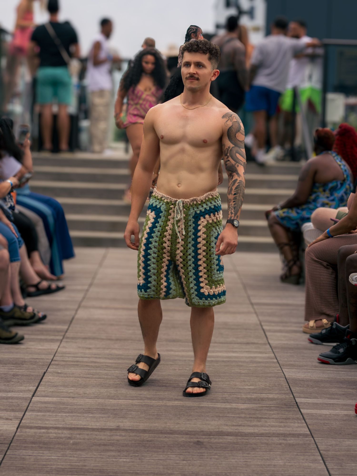 Shirtless tattooed male model in crochet shorts and sandals walks outdoor fashion runway.