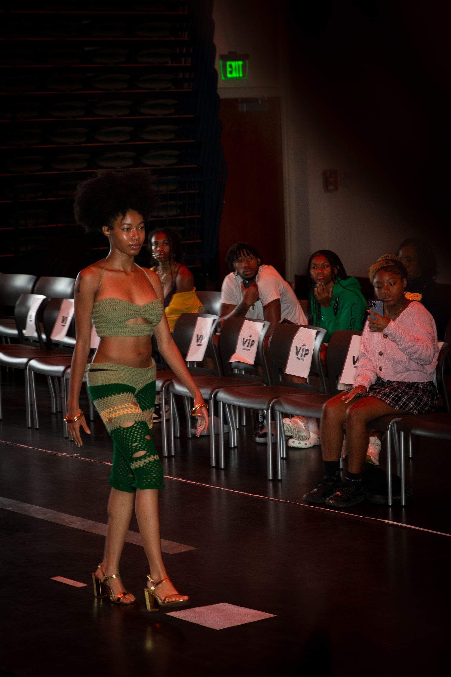 A model in a green crop top and patterned skirt walks the runway at an indoor fashion show.