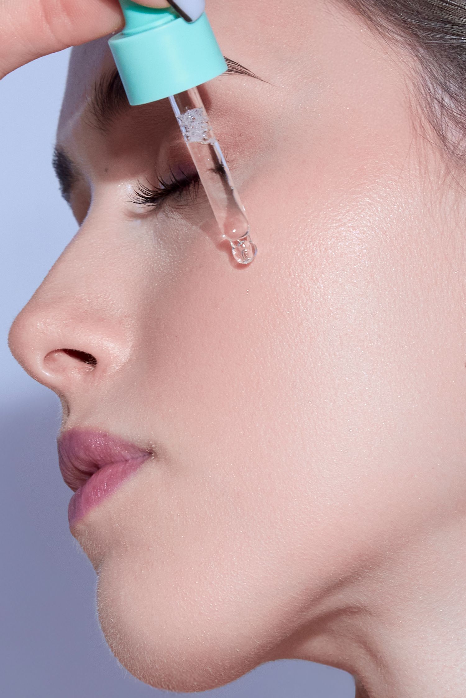 Woman applying clear serum drops to her face using a teal dropper for skincare routine.