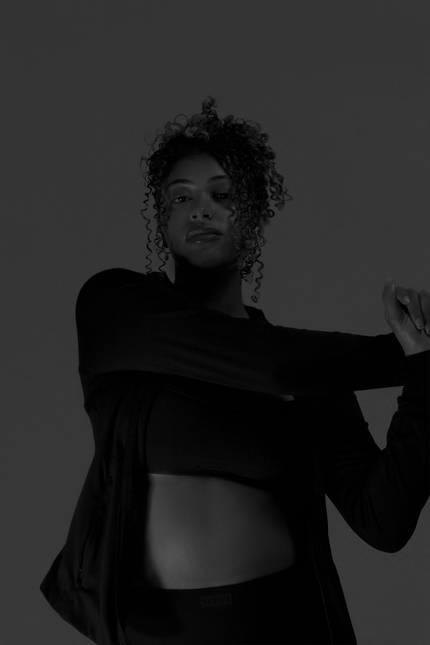 Dark moody black and white portrait of a curly-haired woman in athletic wear posing in studio.