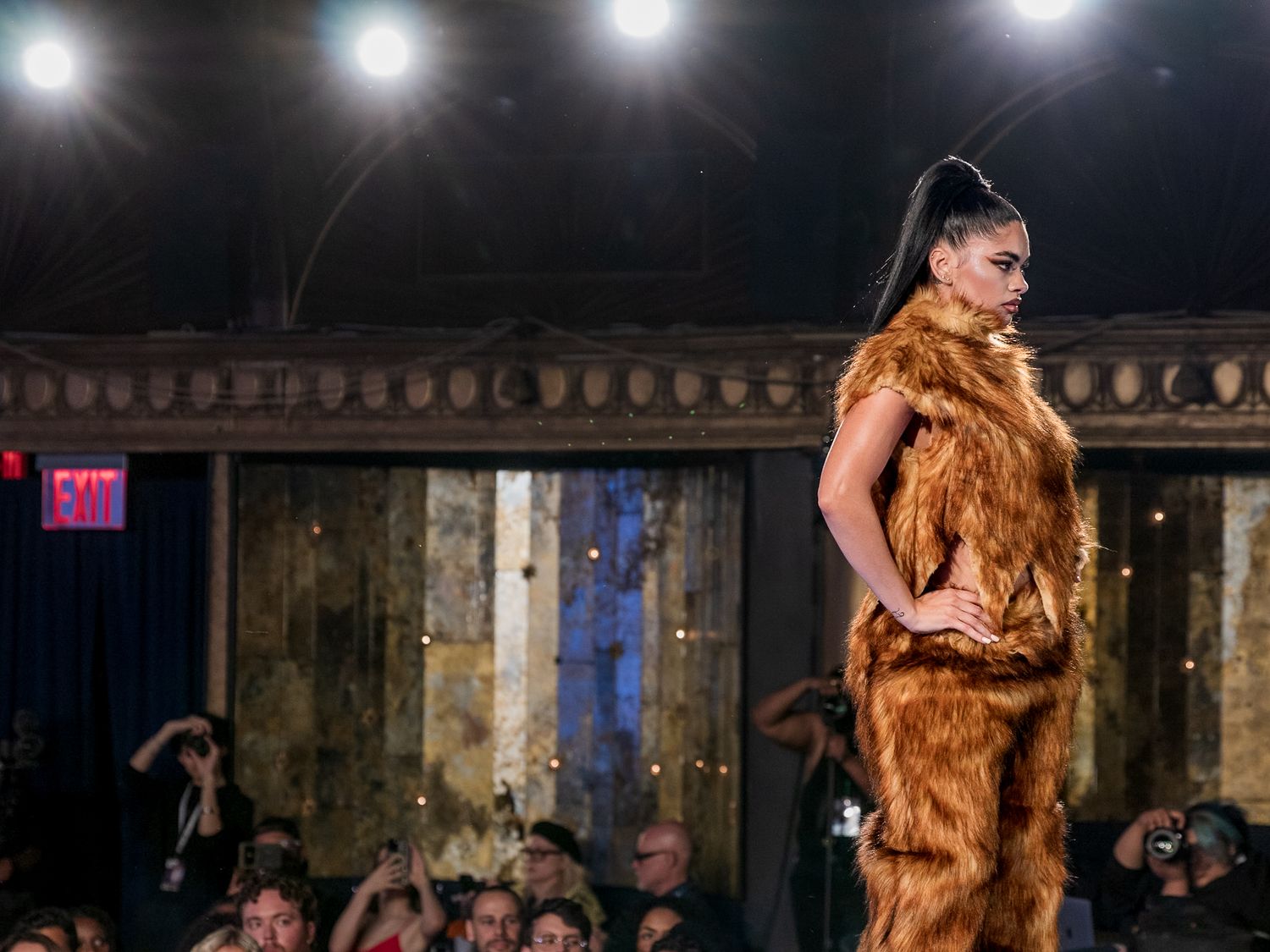 Model walks runway in full golden brown faux fur ensemble at a dimly lit fashion show.