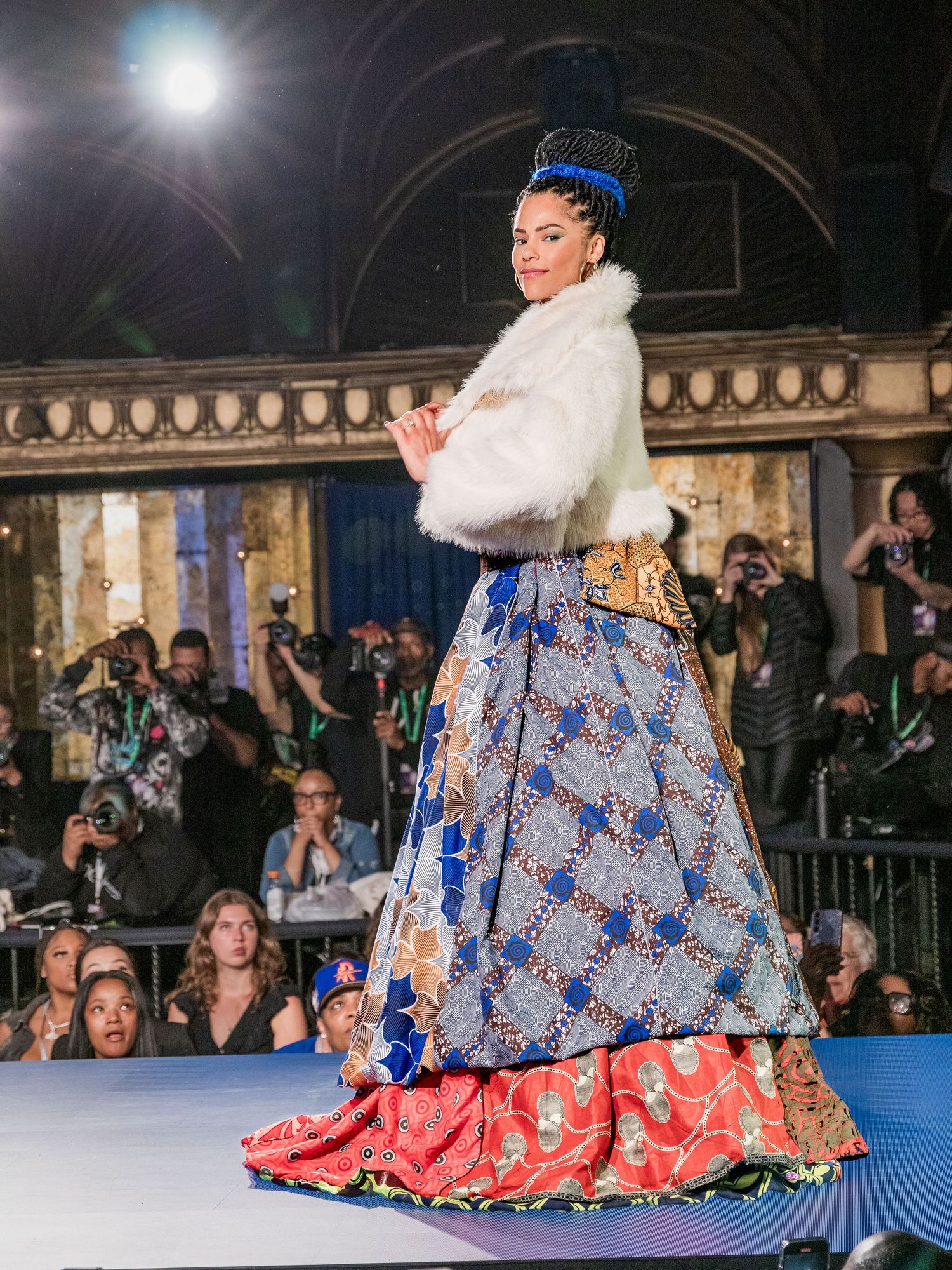 Model poses in layered African print maxi skirt with white fur jacket and decorative blue crown at fashion show.