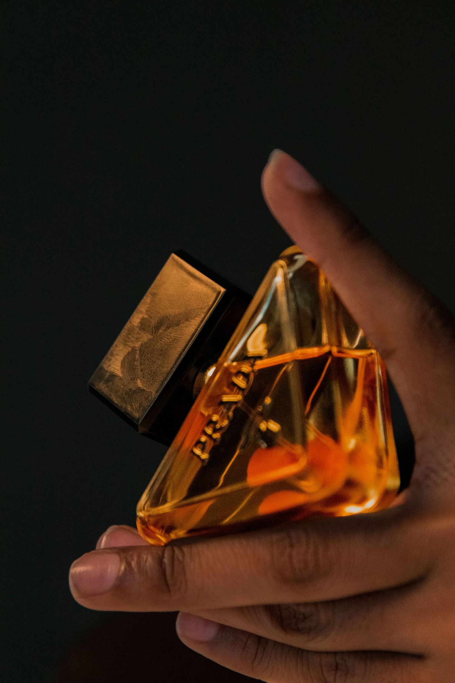Hand holding a luxurious golden Prada perfume bottle against a dark black background.