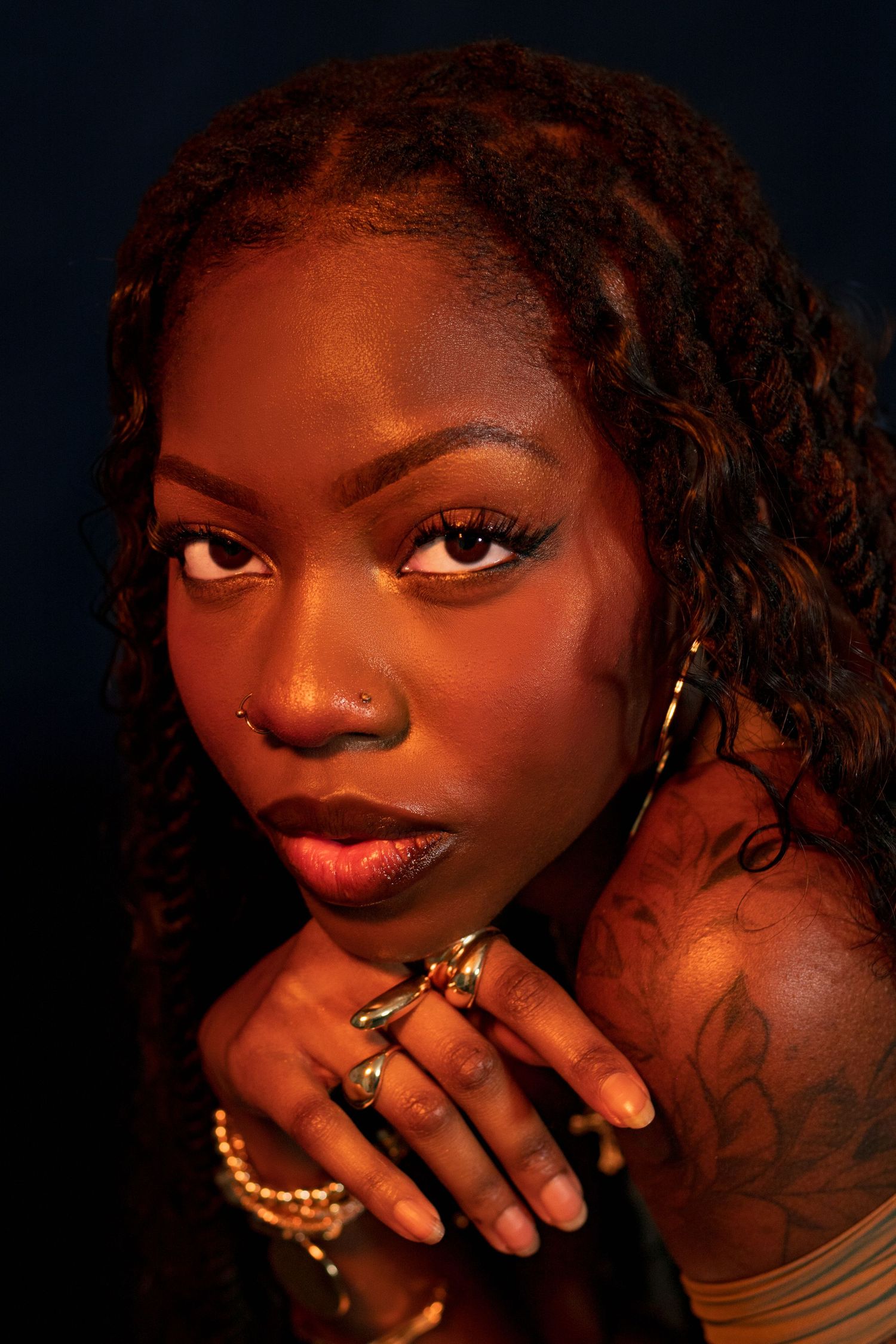 Close-up portrait of a Black woman with tattoos and gold rings resting chin on hands in warm orange studio light.
