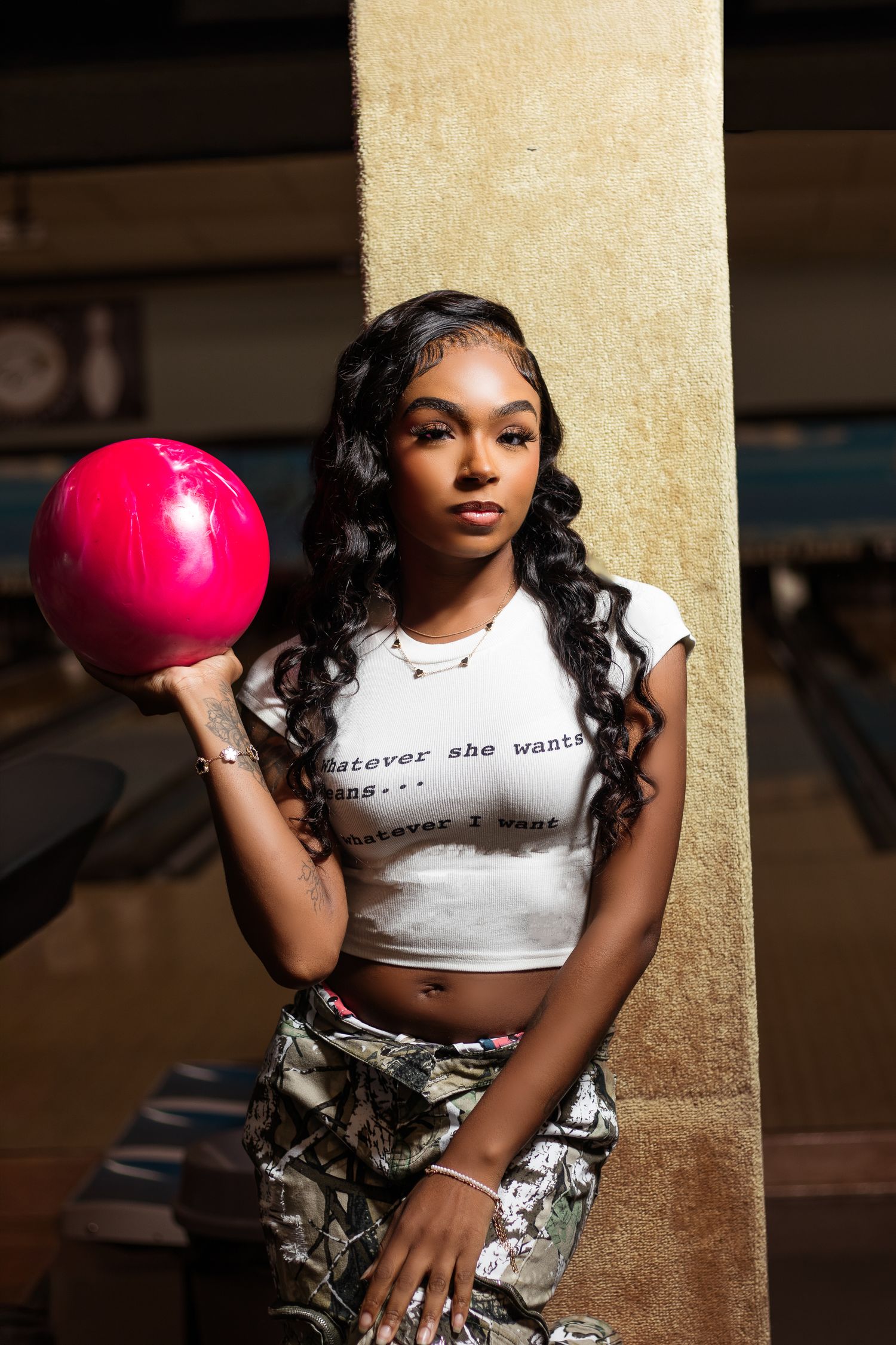 Young Black woman in white crop top holding a pink bowling ball while leaning against a pillar.