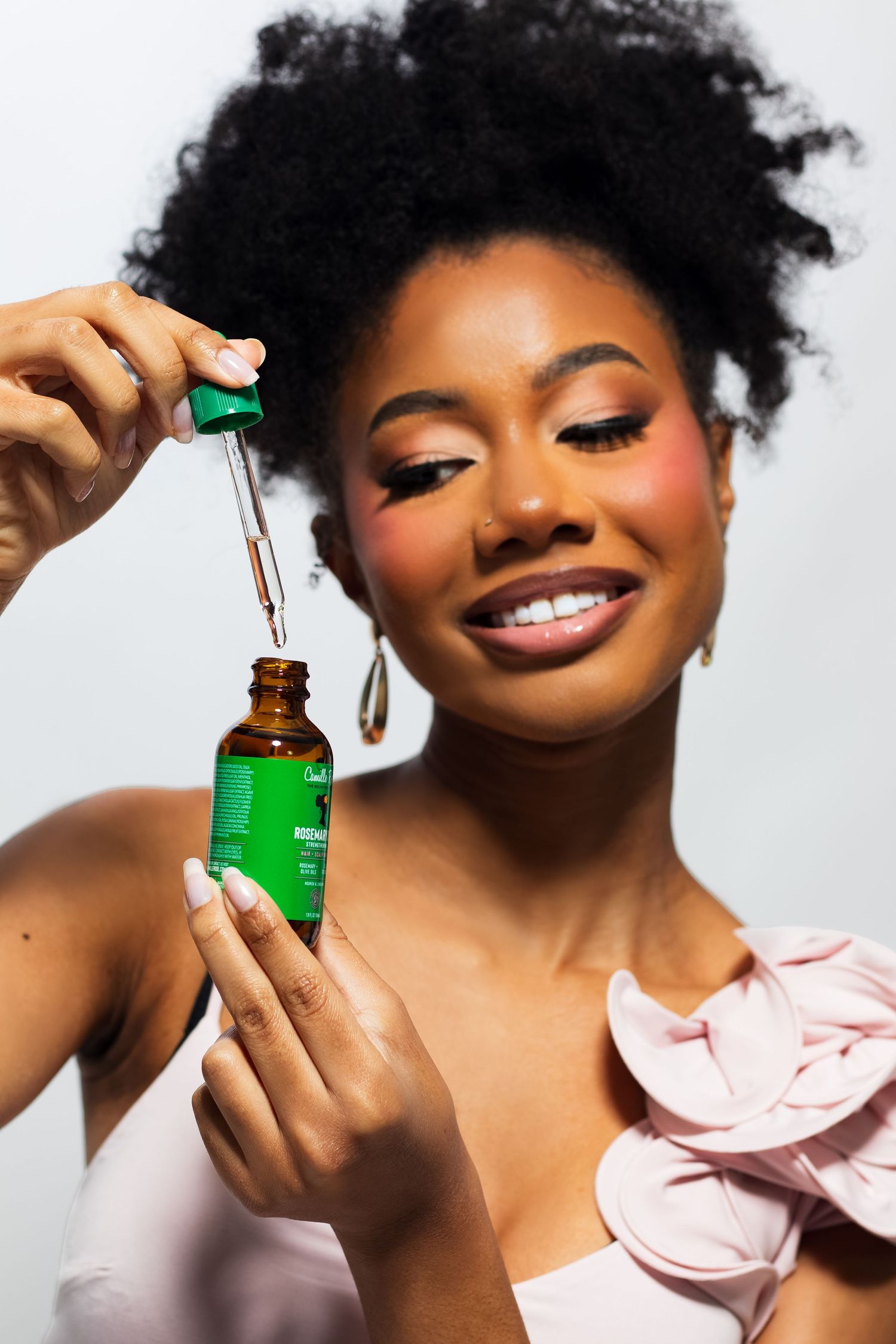 Smiling woman with afro hair using dropper applicator from green herbal serum bottle.