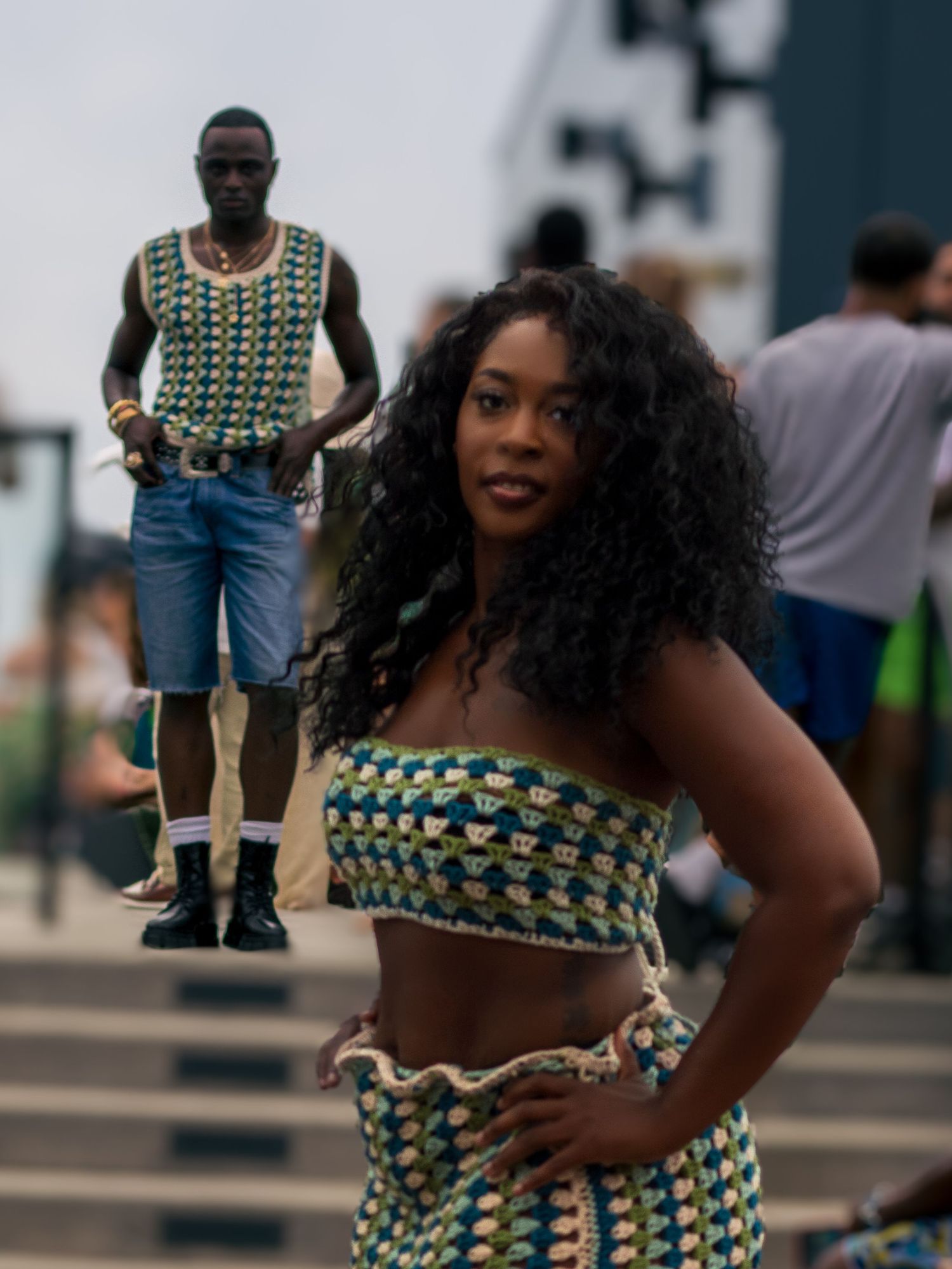 Woman in crochet crop top and skirt poses outdoors with man in matching pattern tank top behind her.