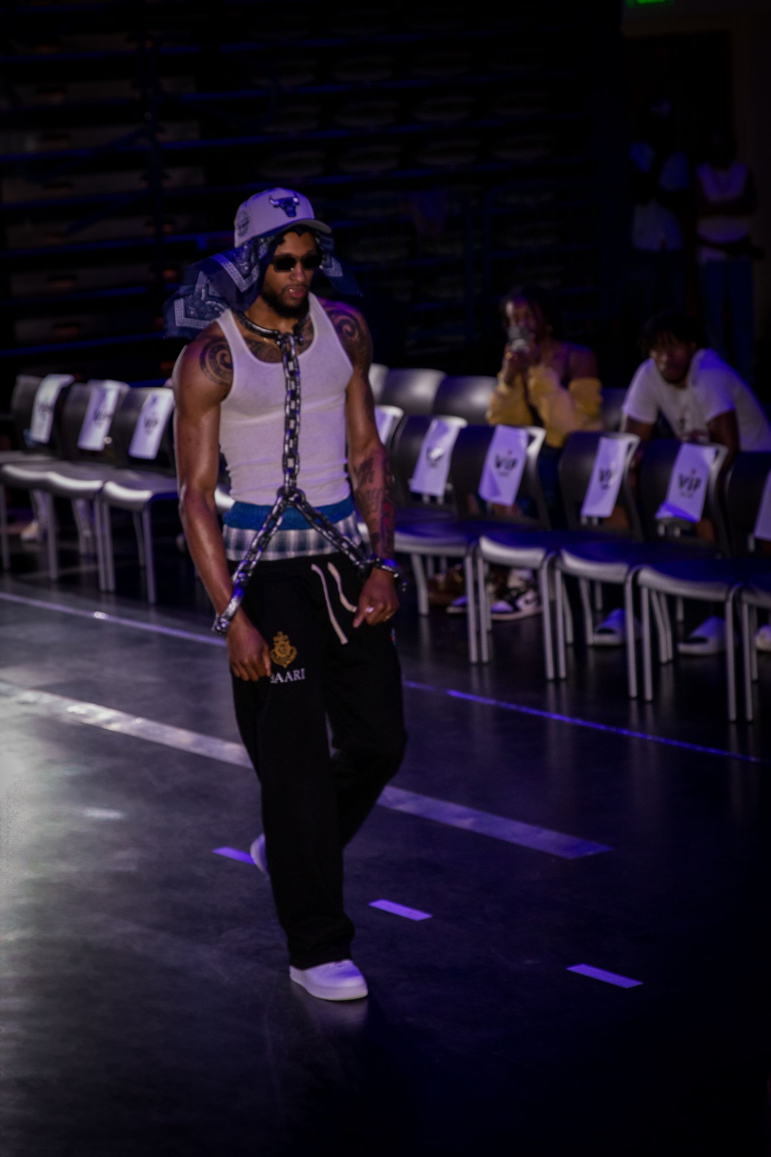 A male model in a white tank top and black pants accessorized with chains walks an indoor runway.