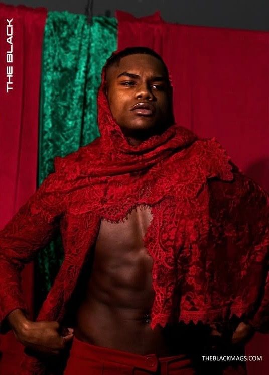 A muscular man poses confidently in an open red lace jacket and matching head wrap against a draped backdrop.