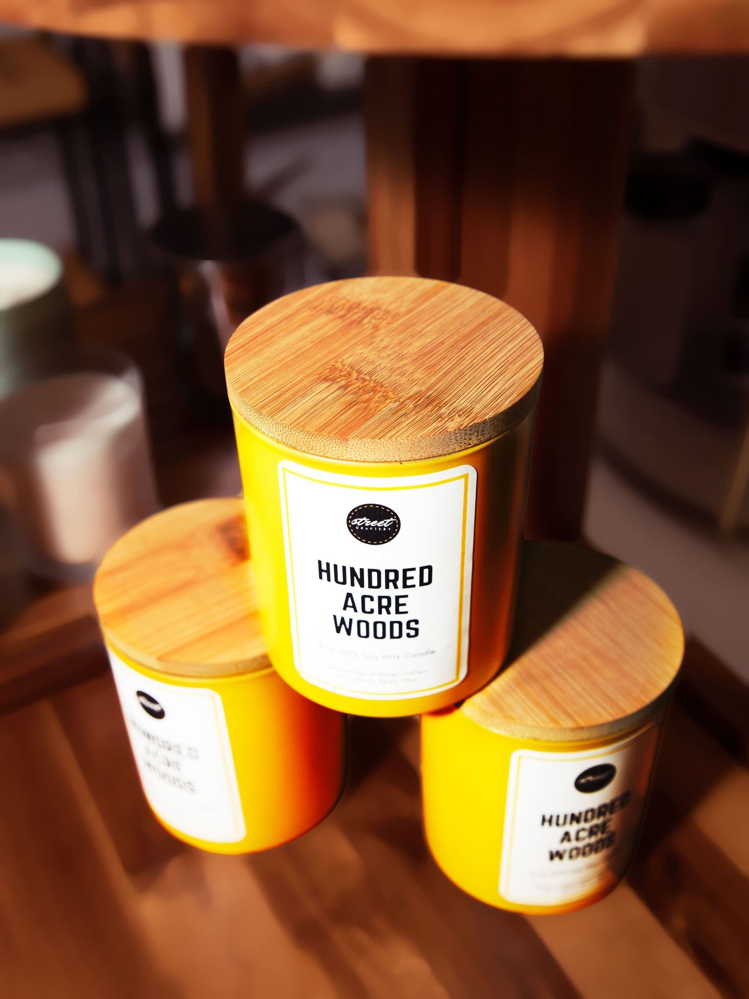 Three yellow Hundred Acre Woods scented candles with bamboo lids displayed on a wooden surface.