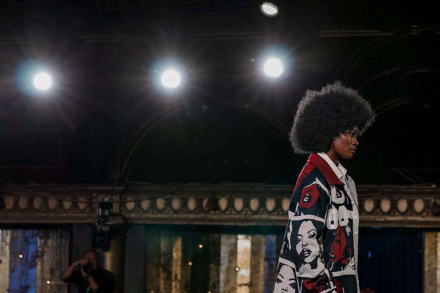 Model with natural afro hair showcases a graphic black and white printed coat under dramatic runway lighting.