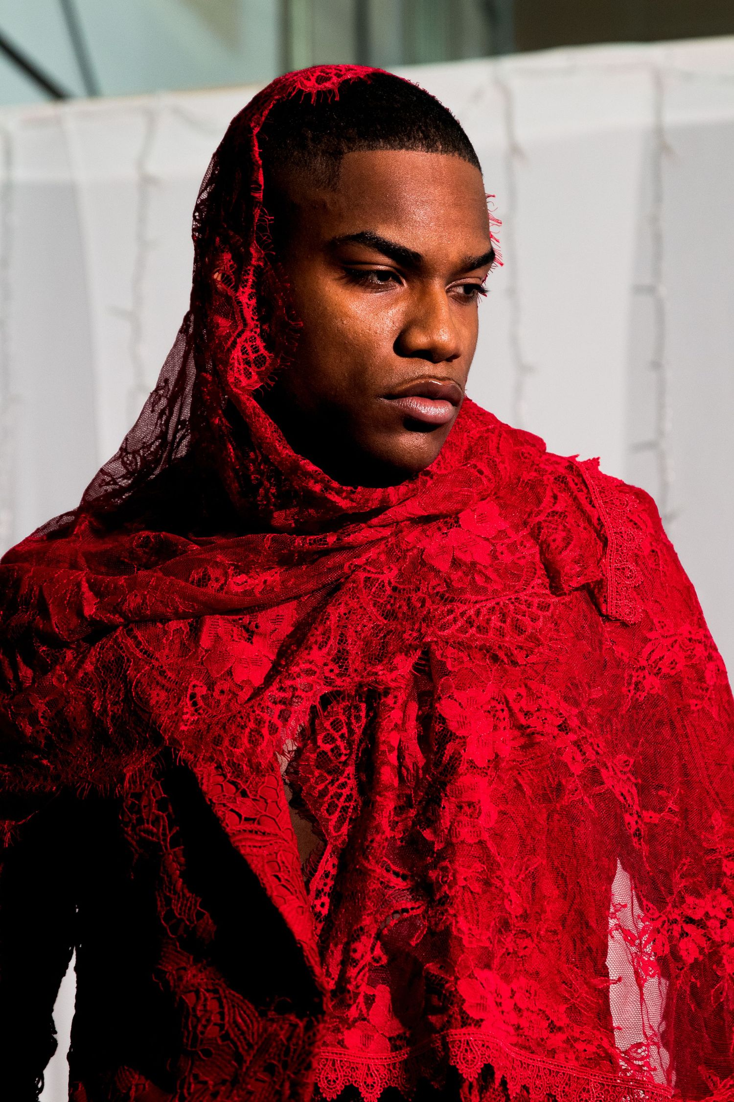 Young man draped in ornate red lace fabric covering his head and shoulders in a fashion portrait.