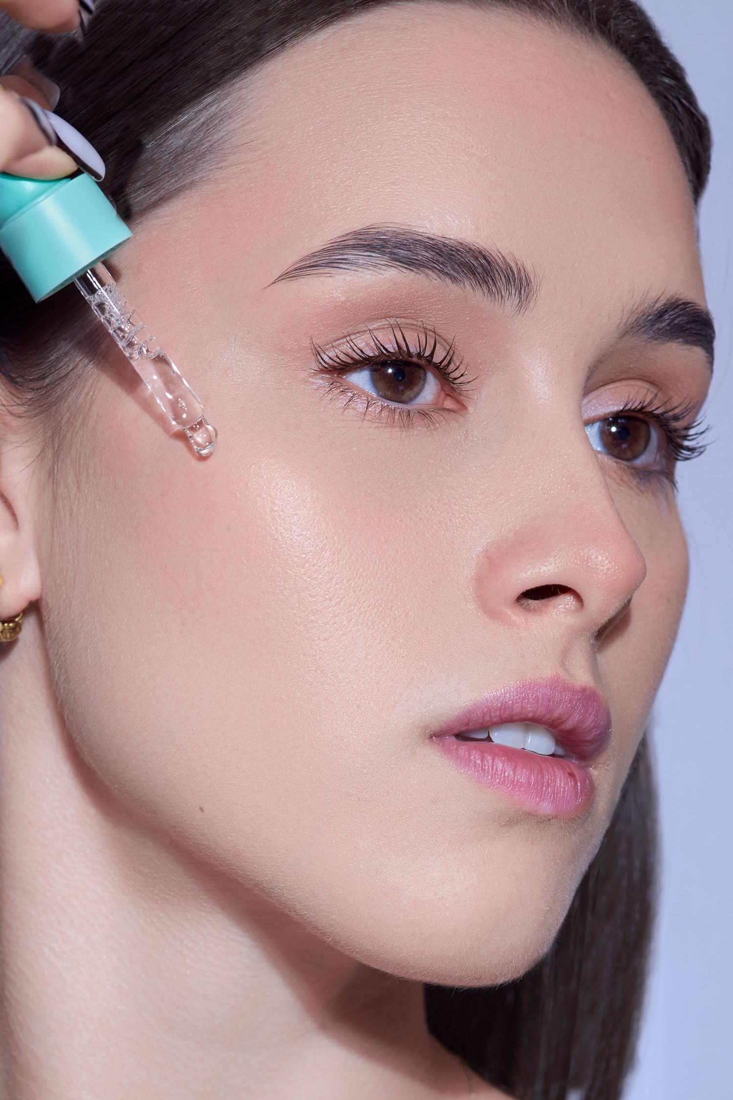 Young woman applying clear serum with a dropper to her glowing skin, showcasing a radiant complexion.