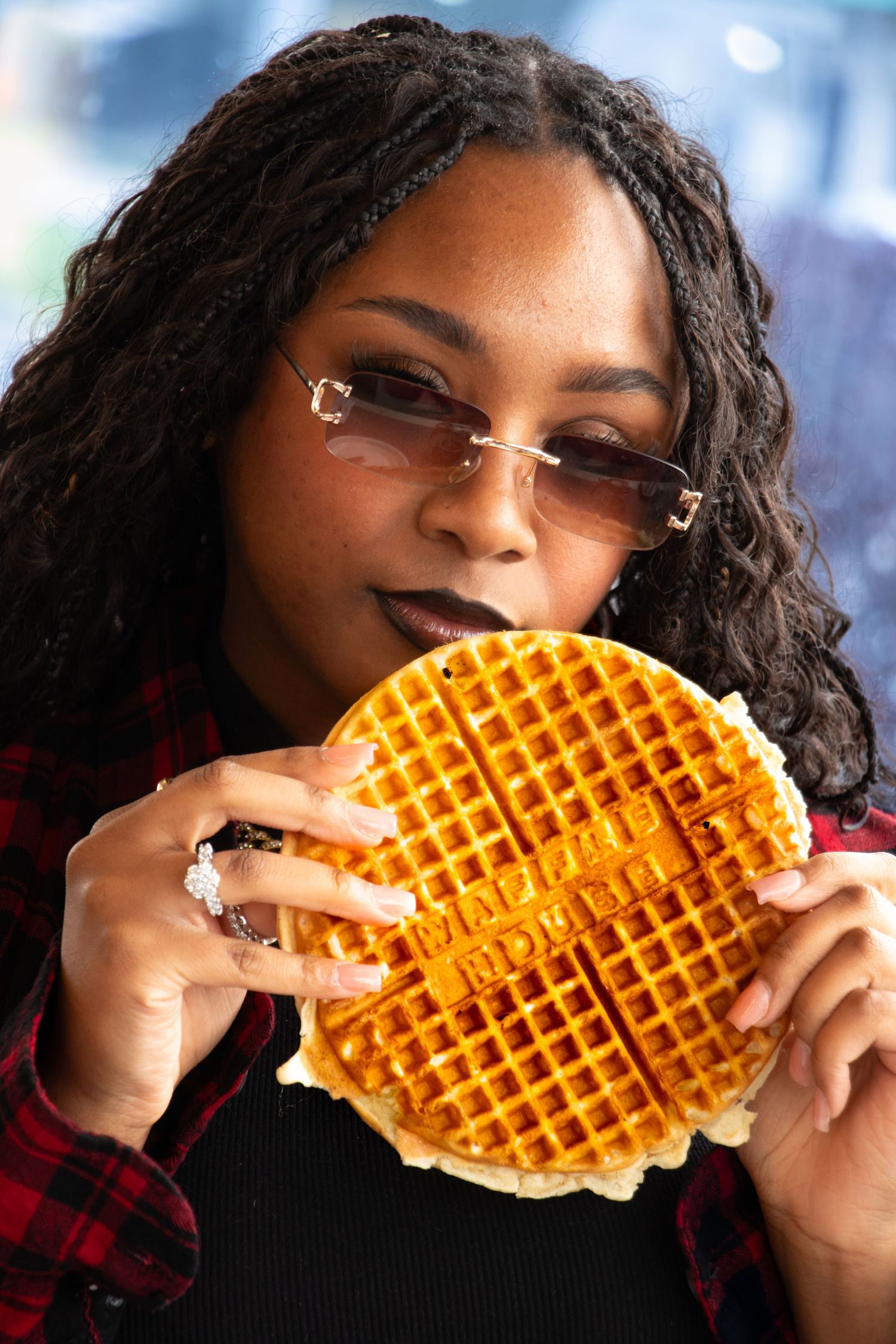 Young woman with braids and tinted glasses holds large branded waffle close to her face.
