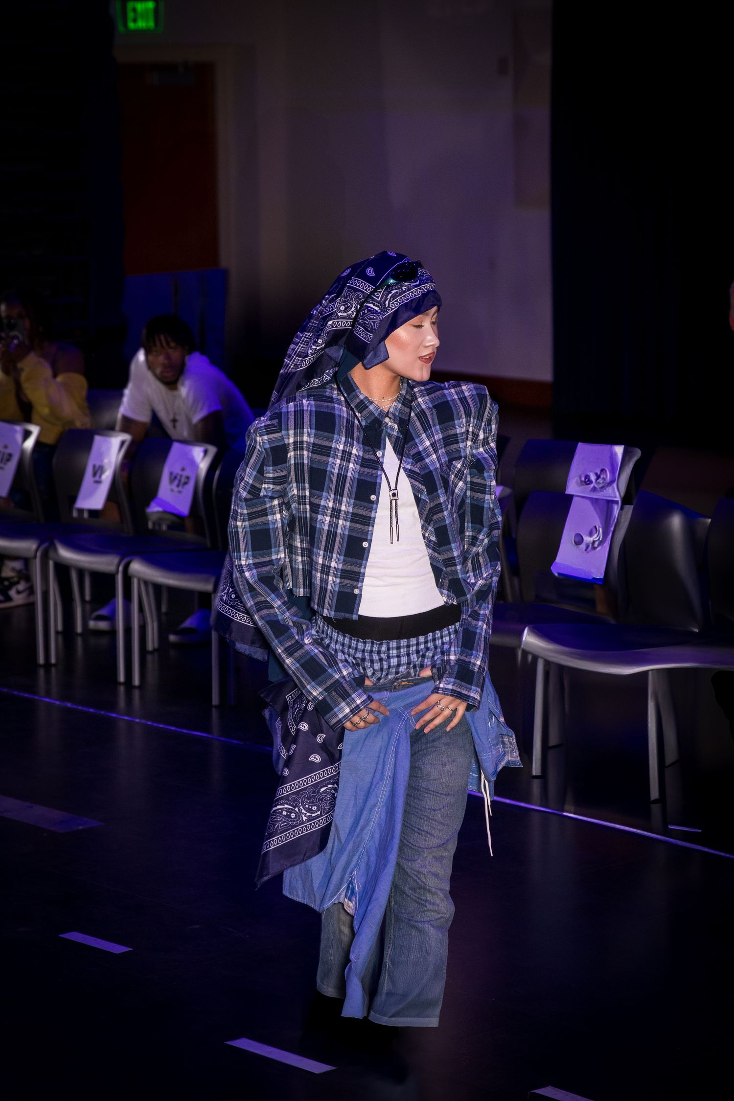 A model in a plaid jacket, jeans, and bandana headwrap smiles on an indoor fashion runway.