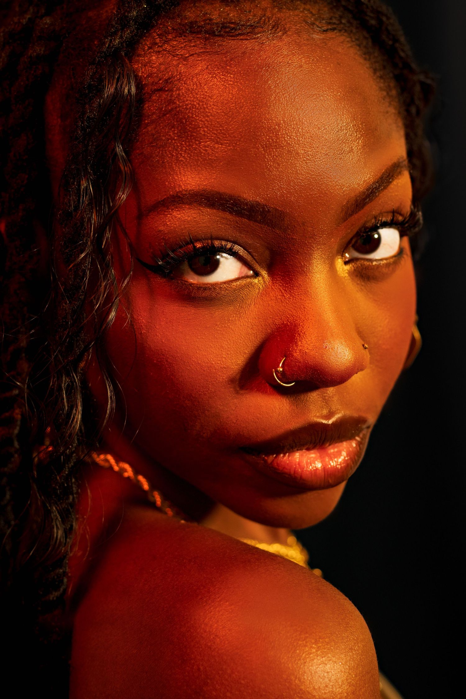Close-up portrait of a Black woman with nose ring and yellow eye makeup in dramatic warm orange studio lighting.