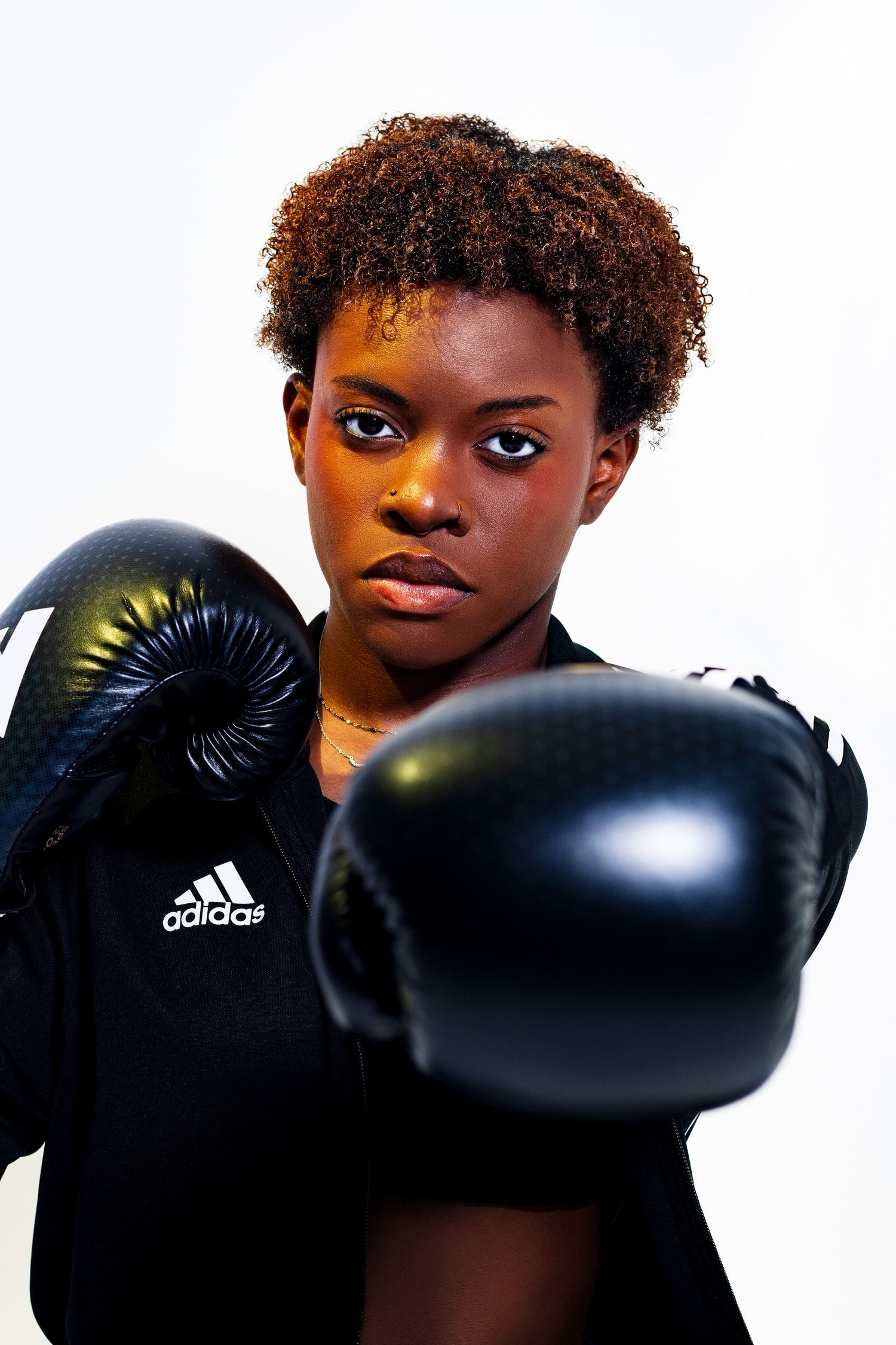 Determined young Black female boxer in Adidas jacket in fighting stance with black boxing gloves raised.