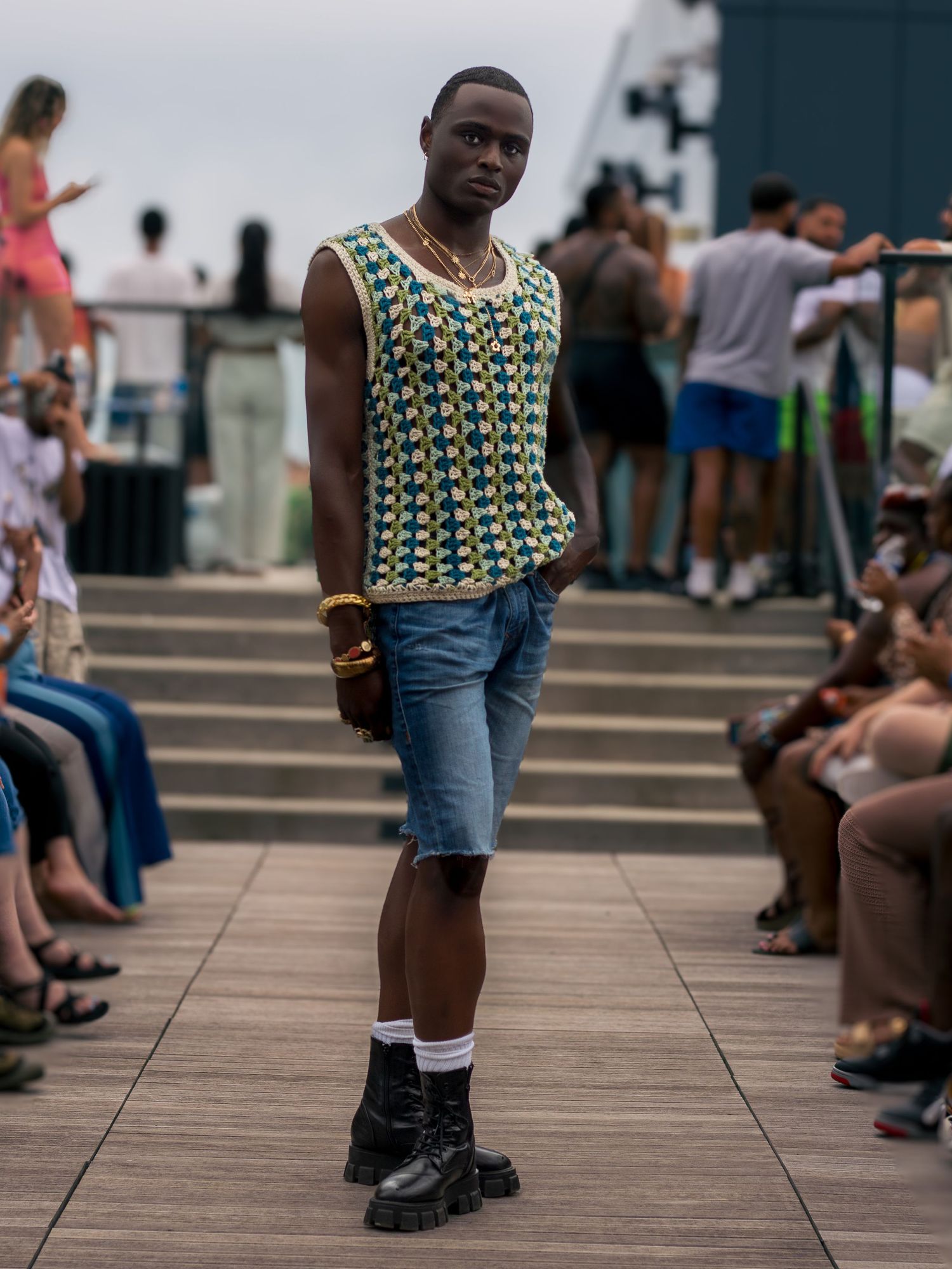 Male model in crochet tank top, denim shorts, and black boots on outdoor runway.