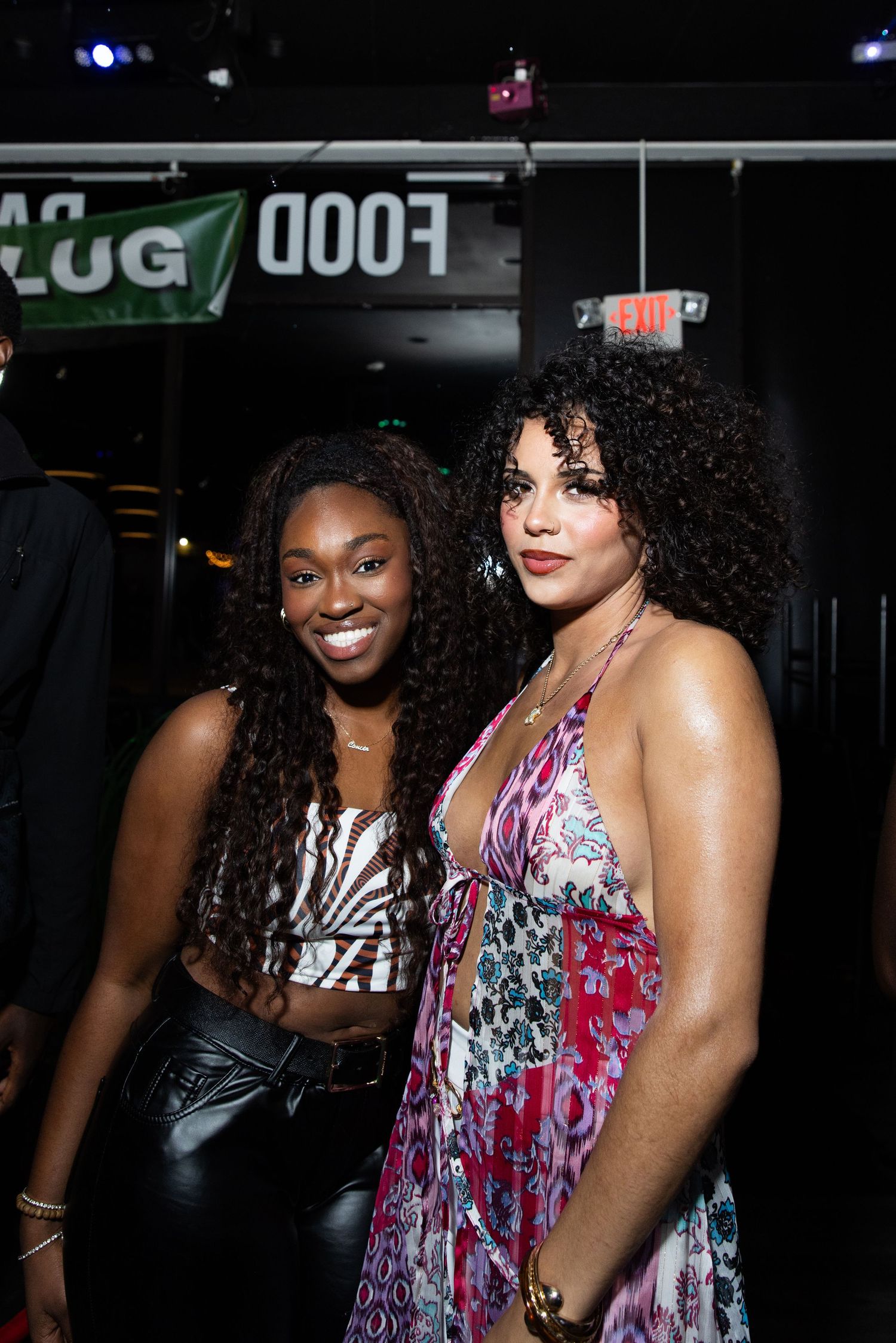 Two women smile together at a nightclub near a food sign.