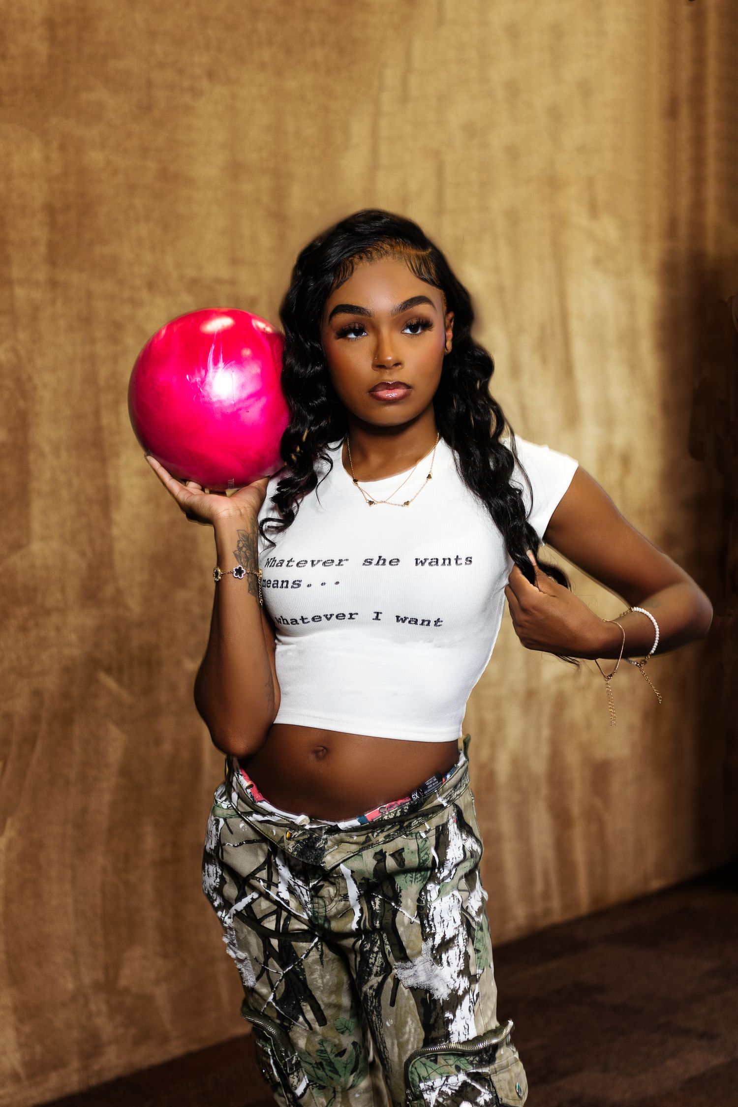 Young Black woman in white crop top and camo pants posing with a pink bowling ball indoors.