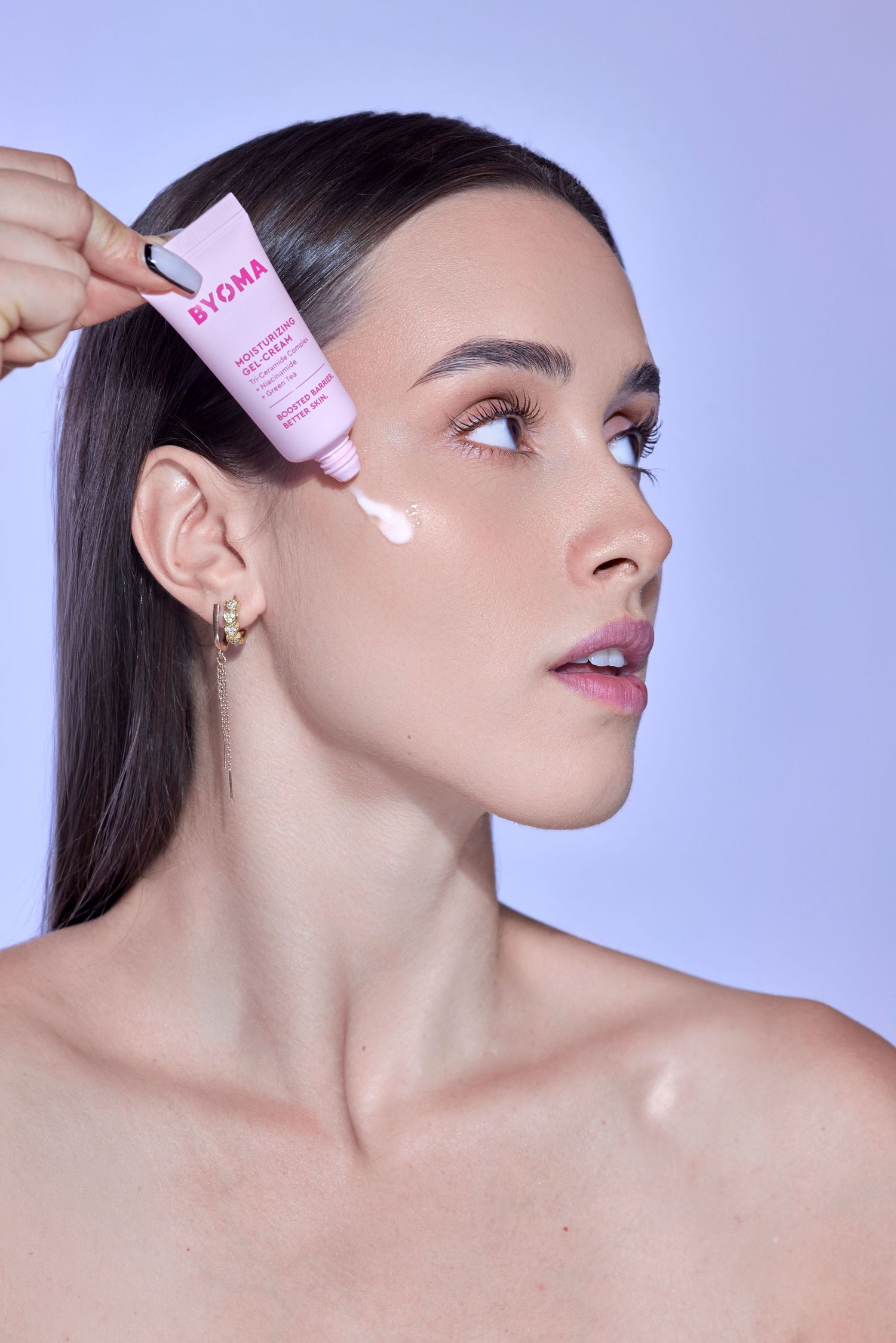 Woman applying Byoma moisturizing cream from a pink tube to her face against a lavender background.