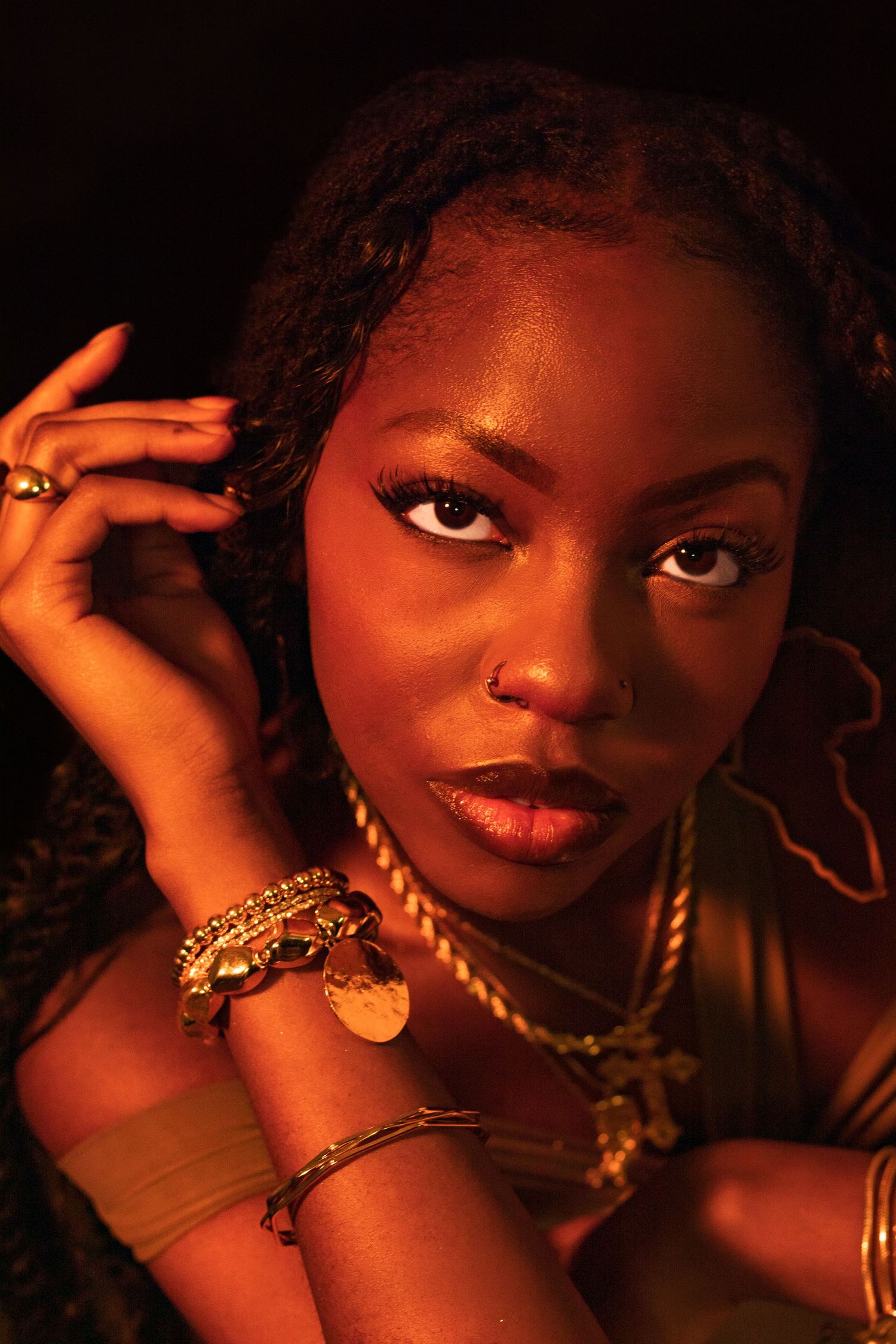 Close-up portrait of a Black woman with gold jewelry and bracelets gazing at camera in warm golden studio lighting.
