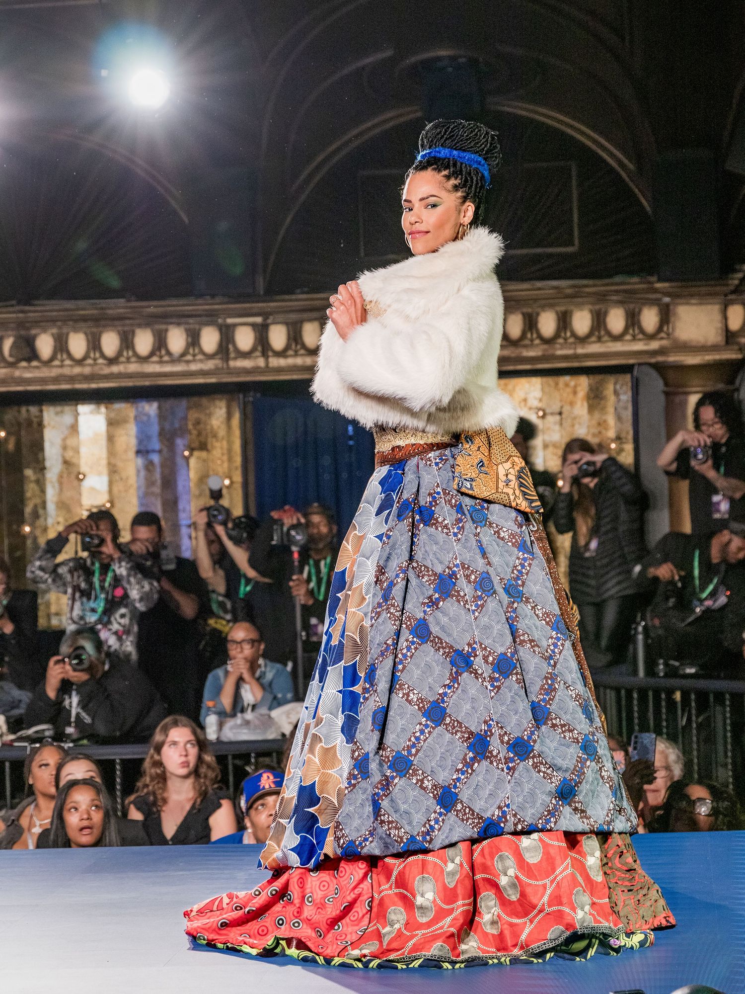 Model walks runway wearing layered colorful African print skirt, white fur shrug, and ornate blue crown headpiece.