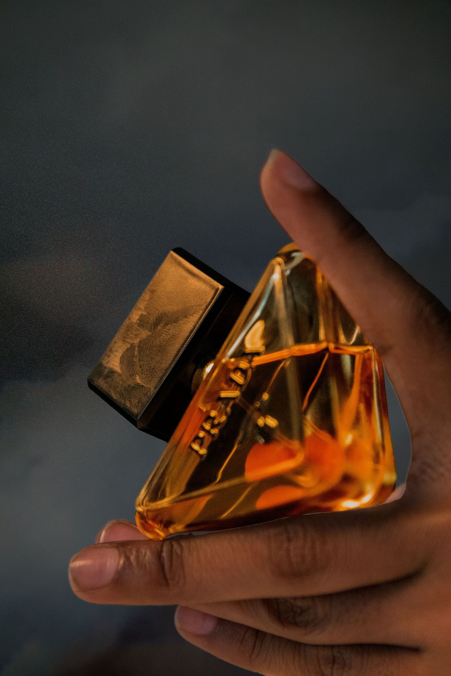 Hand holding a triangular amber glass perfume bottle with golden cap against a dark background.