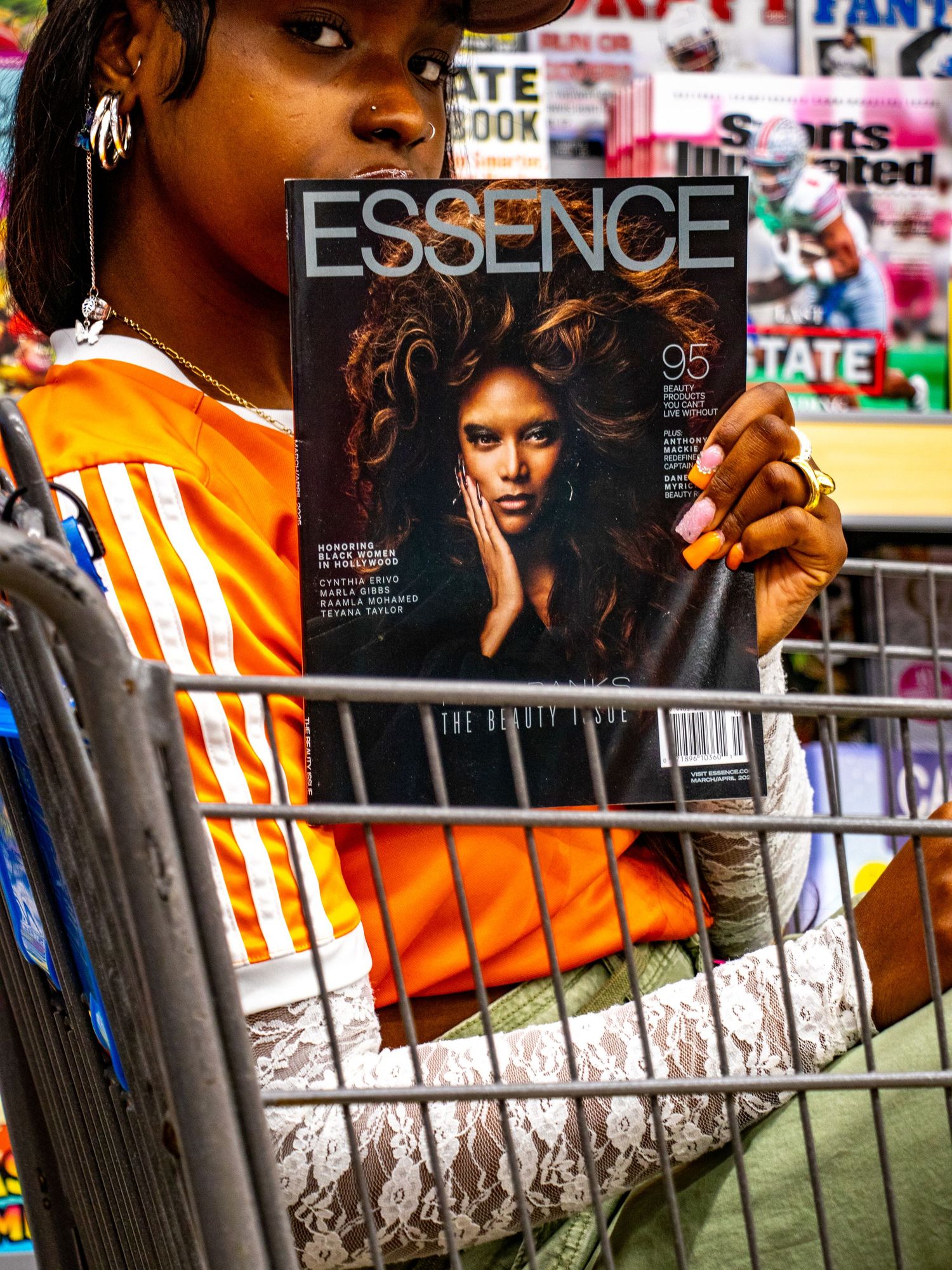 Young woman in orange jersey sits in shopping cart holding Essence Beauty Issue magazine in store.