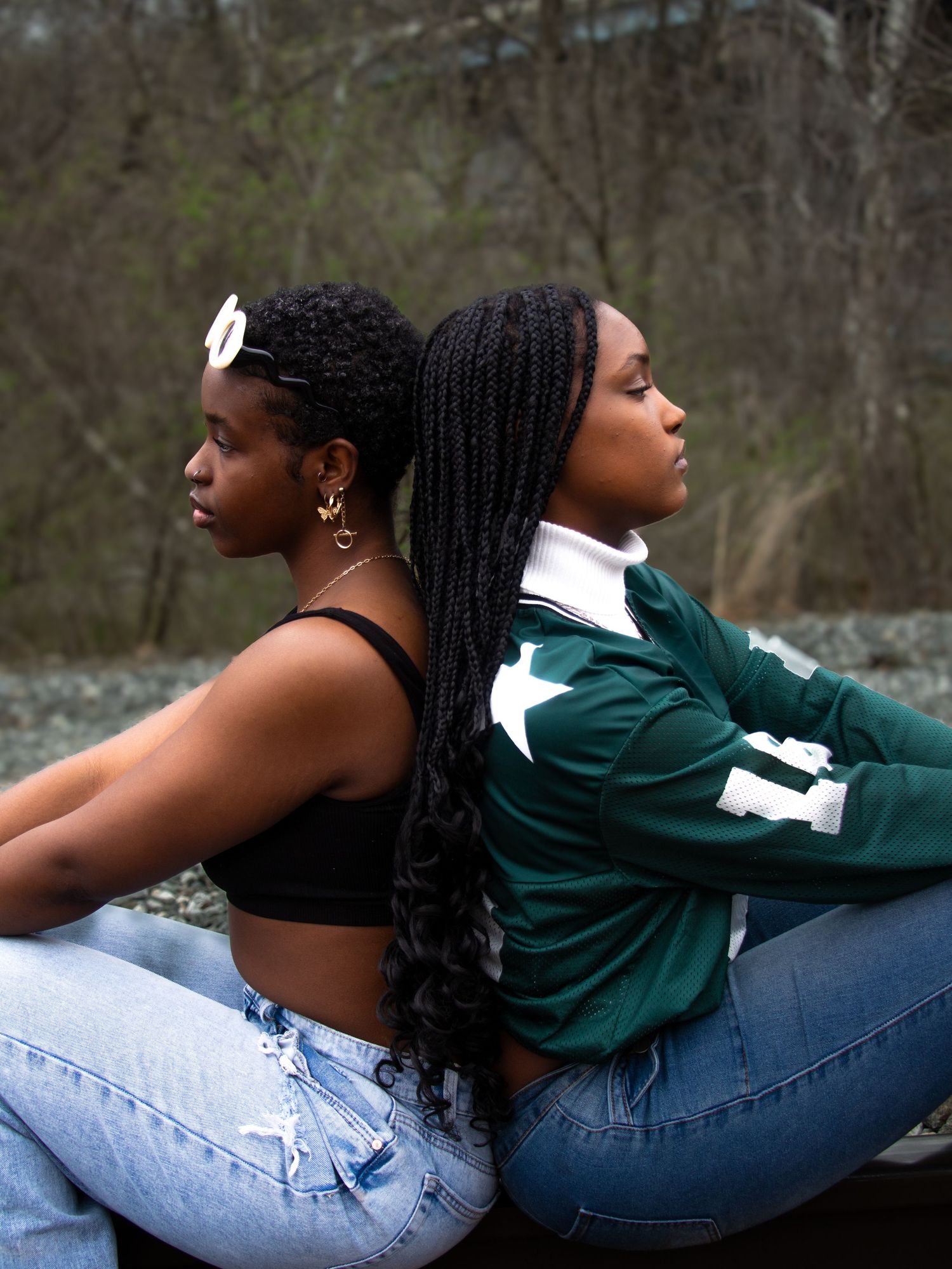 Two young women sit back-to-back outdoors, one in a black crop top and one in a green San Francisco jersey with blue jeans.