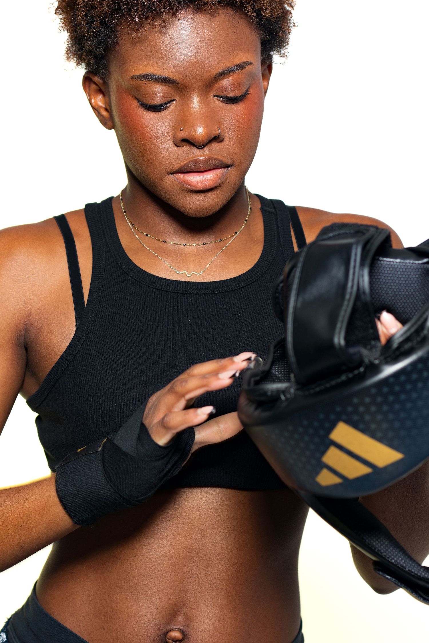 Young Black female boxer putting on Adidas black and gold boxing gloves wearing sports bra.
