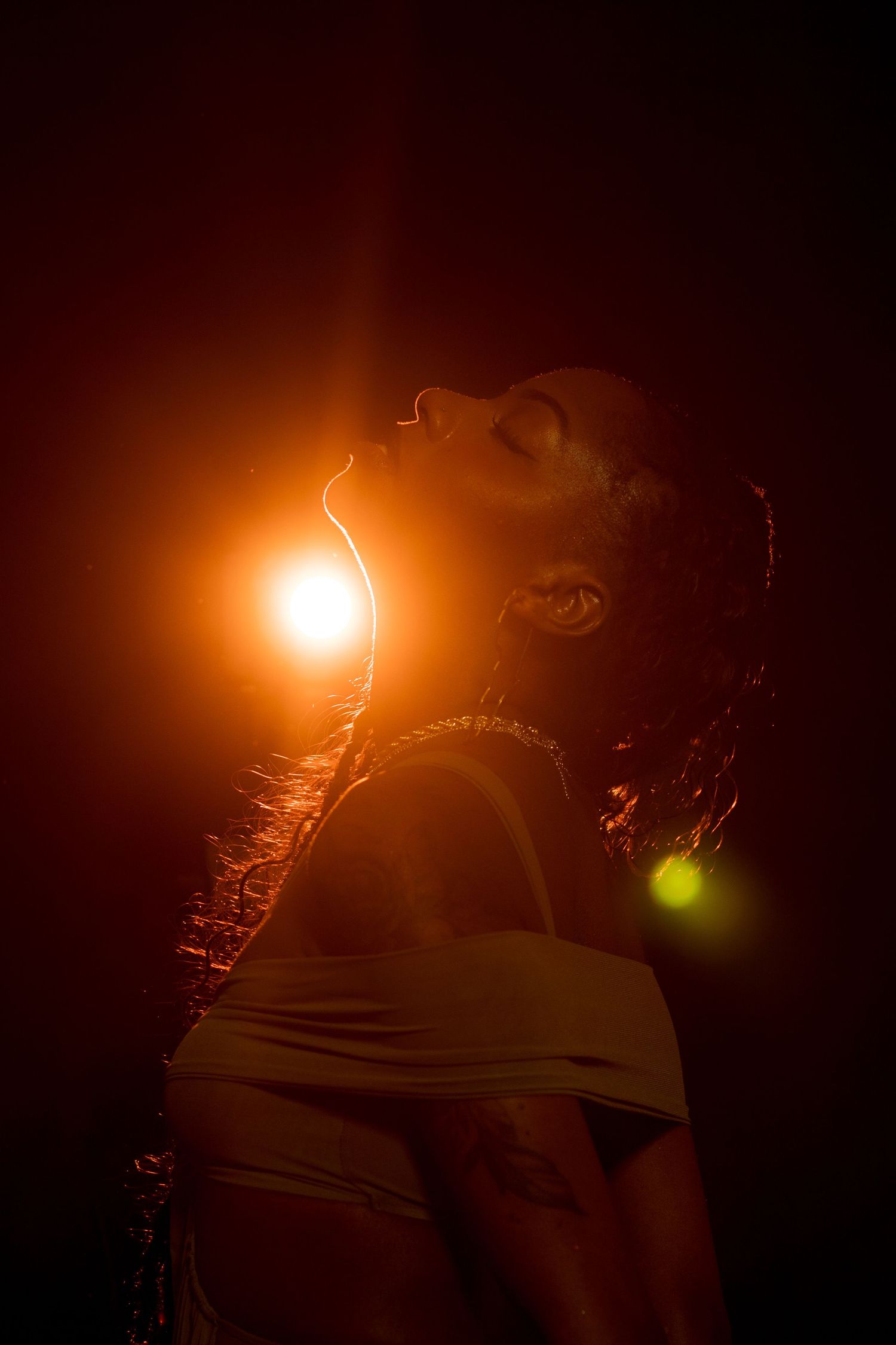 A Black woman tilts her head back dramatically with glowing orange backlight illuminating her profile in a dark studio.