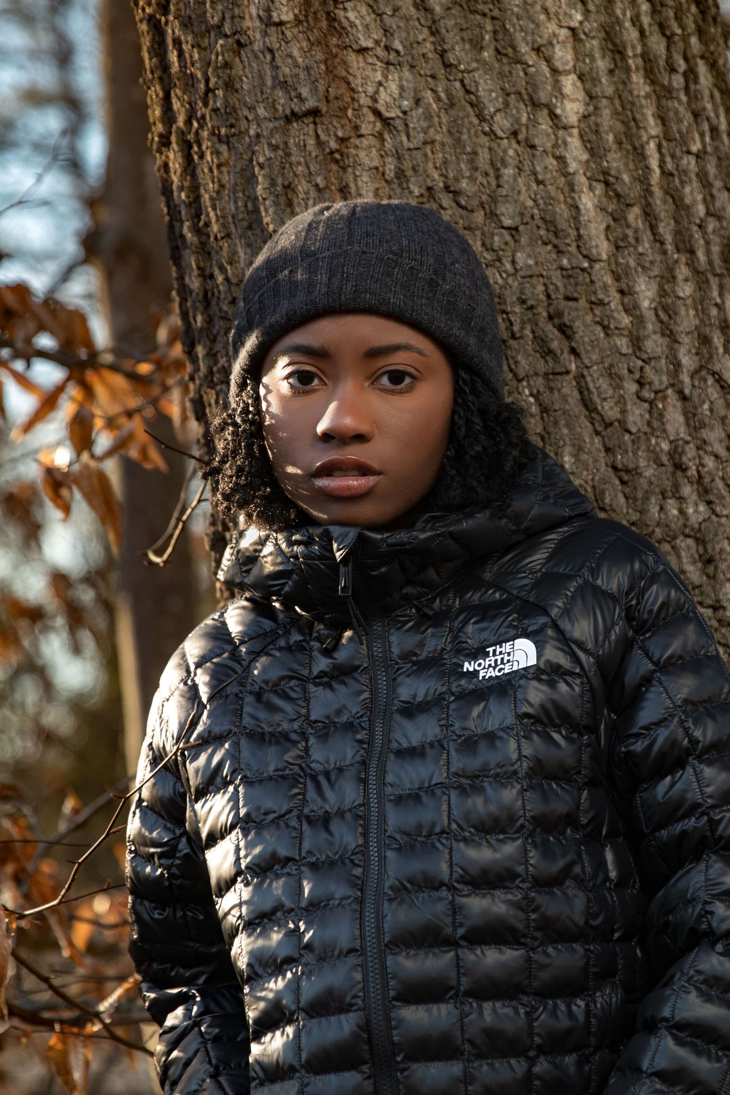 Young woman in black North Face puffer jacket and beanie leaning against tree outdoors.