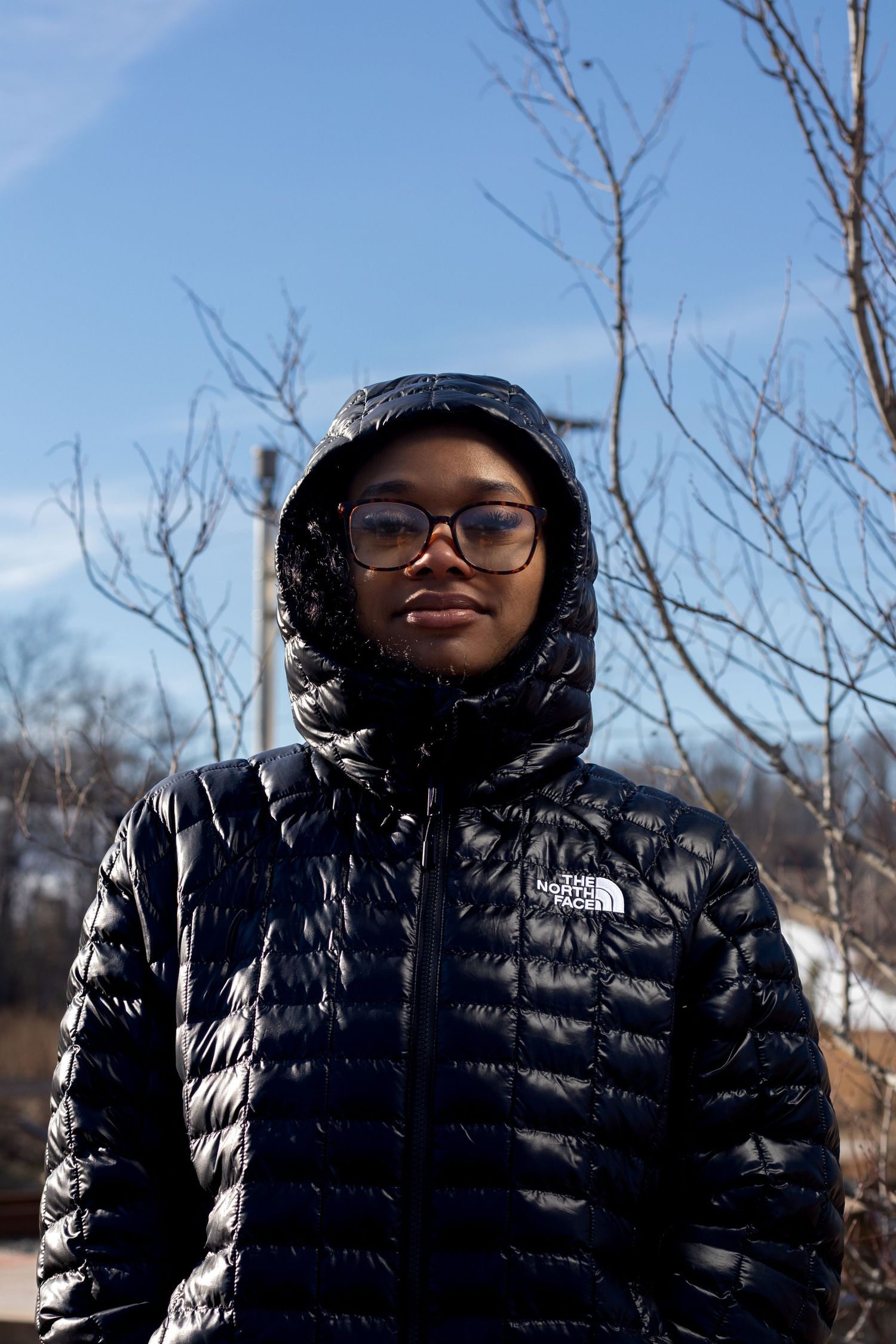 Person wearing glasses and black The North Face hooded puffer jacket against blue sky.
