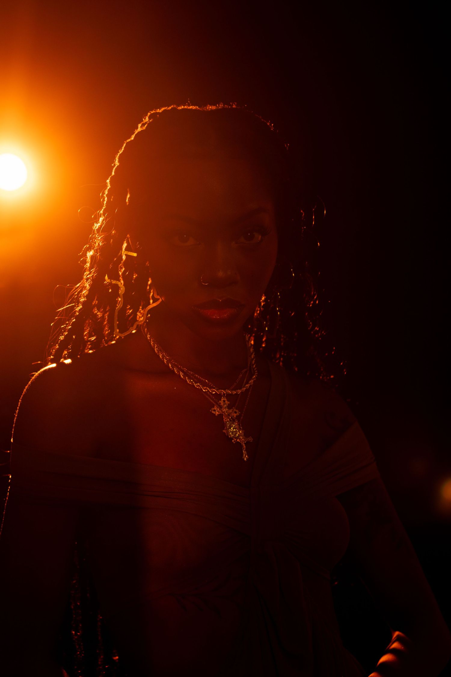 A person with dreadlocks and a cross necklace poses dramatically under warm orange backlit studio lighting.