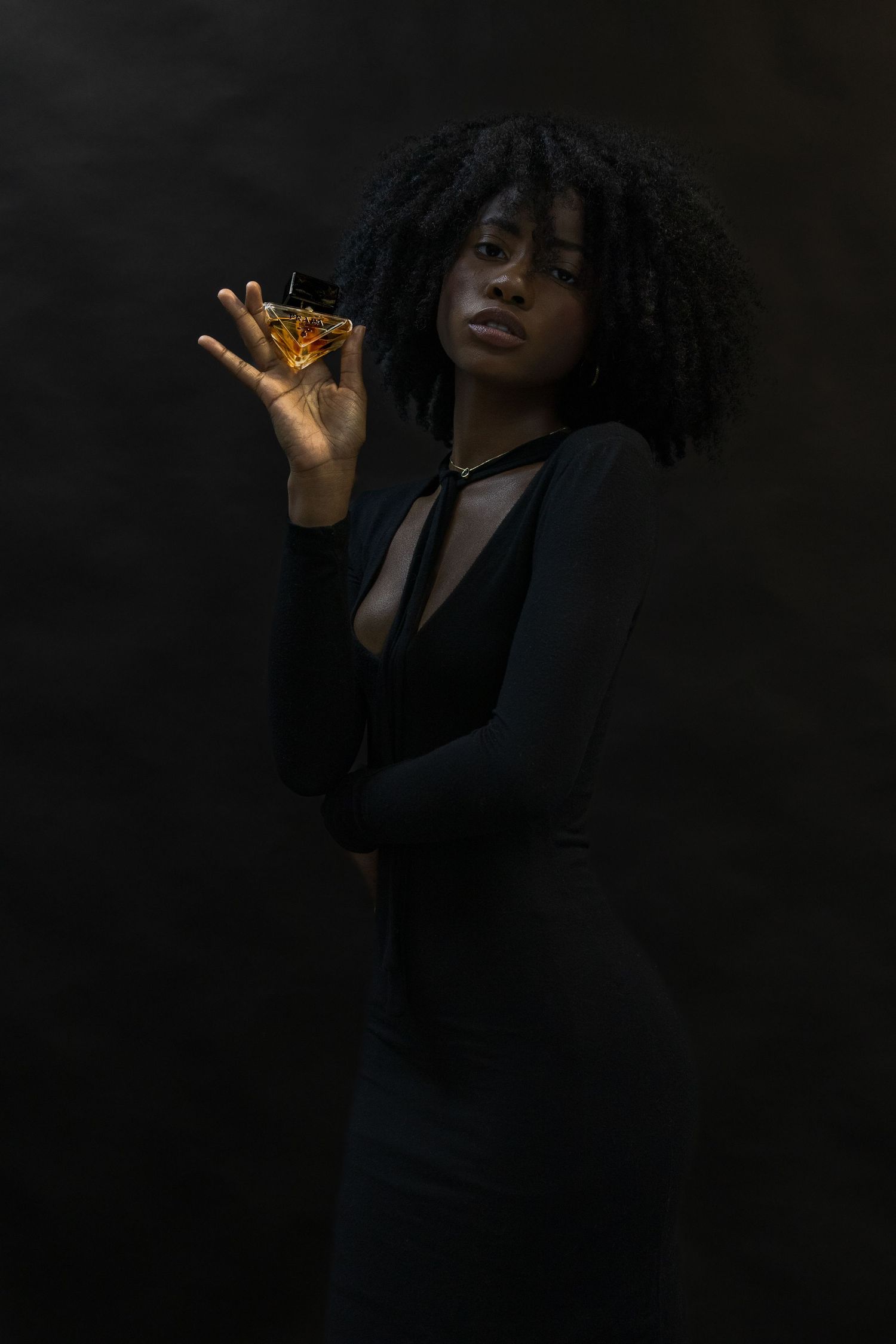 Elegant woman in black dress holding a Prada fragrance bottle in dramatic dark studio lighting.