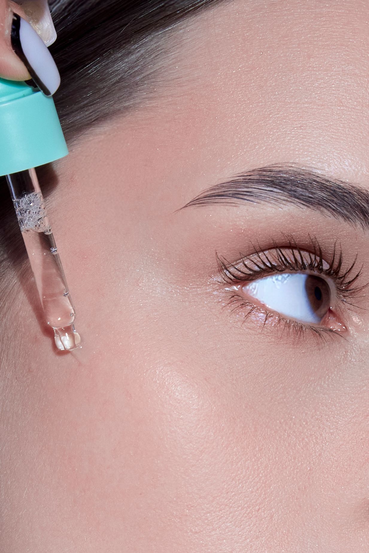 Close-up of a woman applying clear serum drops from a teal dropper bottle near her eye on glowing skin.